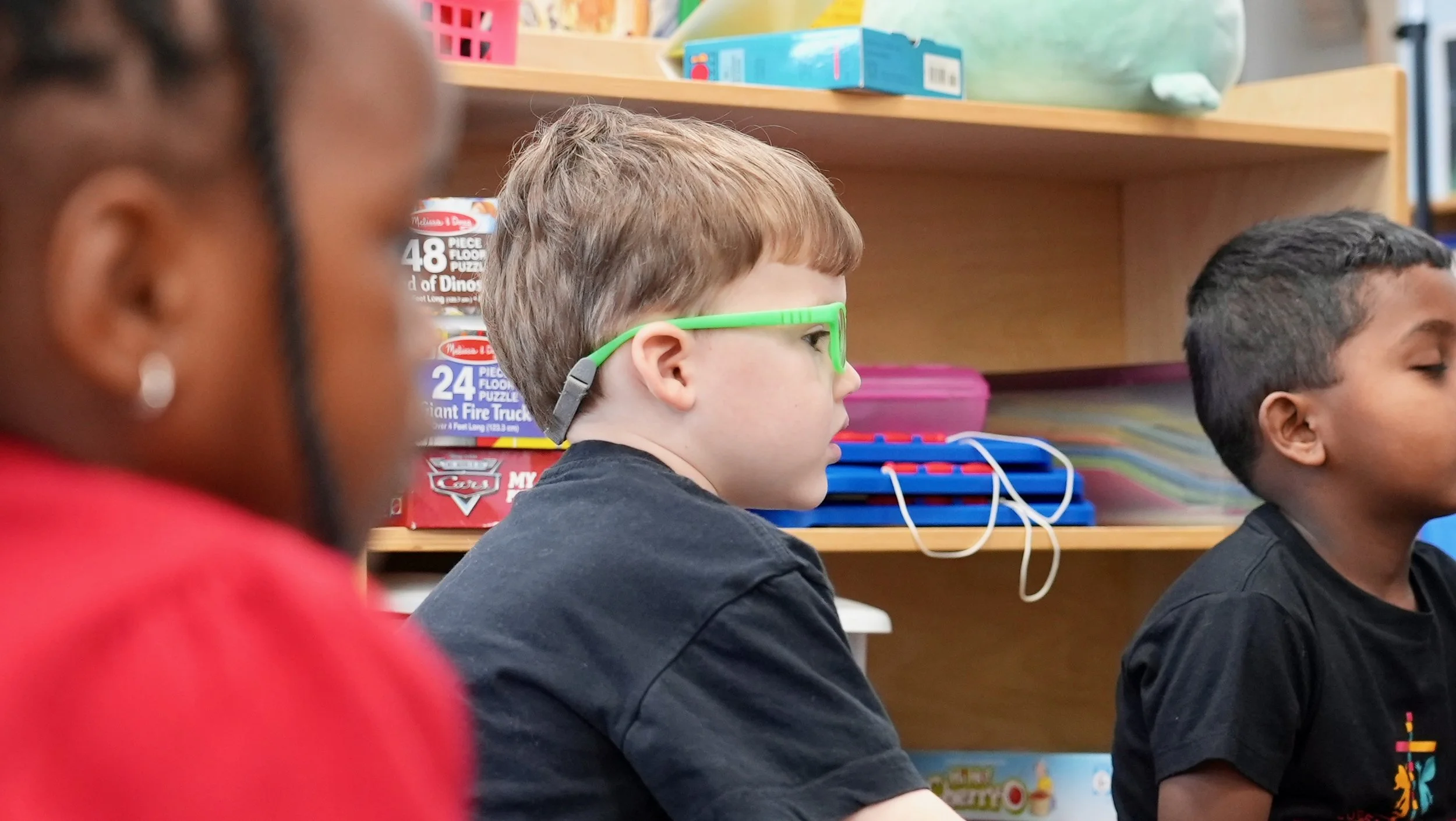 Students at Redeemer Lutheran Preschool in Olathe, Kansas, engaged in a classroom activity, with educational materials and games on shelves in the background.