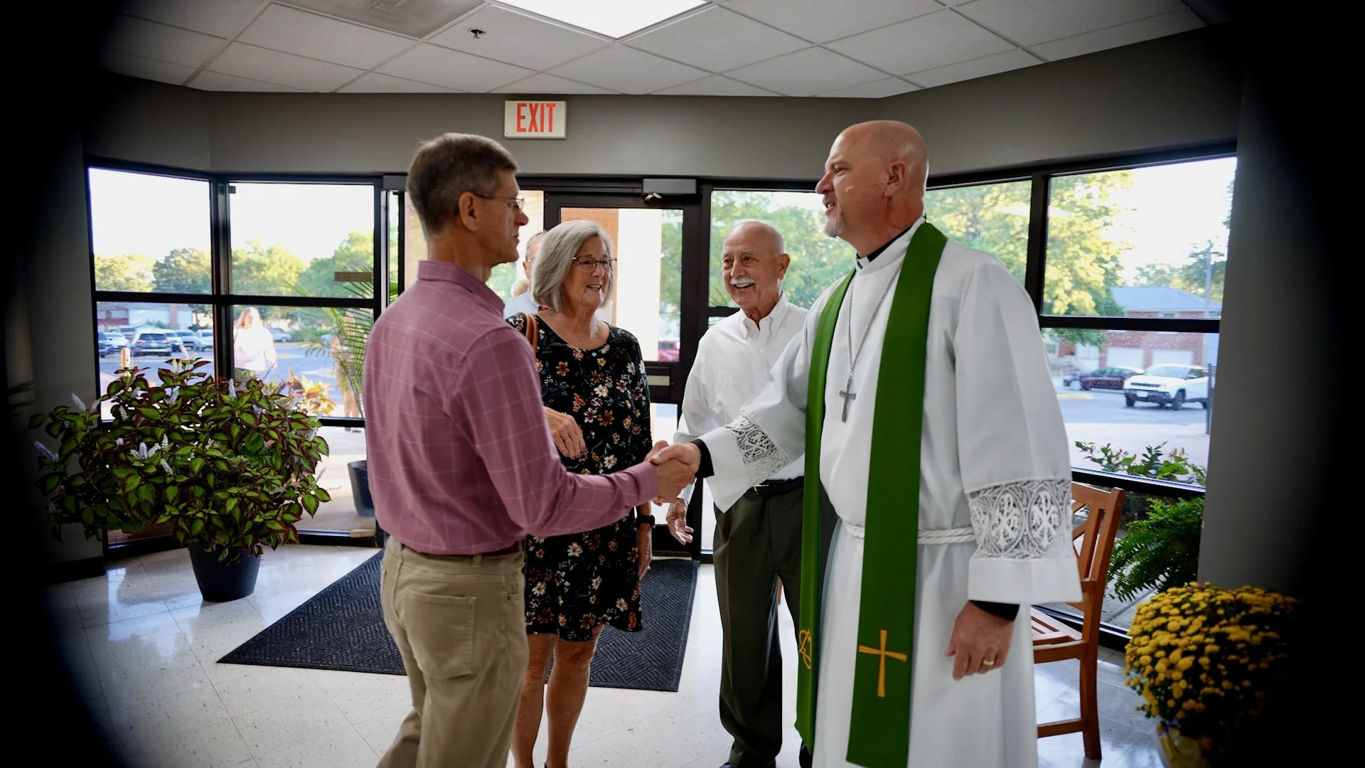 A priest with a green stole shaking hands with a man in a pink shirt inside a church or community center. Two women and an older man are smiling nearby as they watch the interaction.