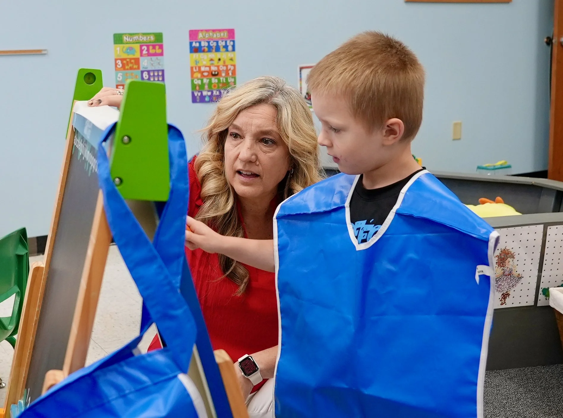 Corrie Klatt, Preschool Director at Redeemer Lutheran Preschool, guiding a young student during a hands-on learning activity at a small classroom easel. Both are engaged in creative play and early childhood education.