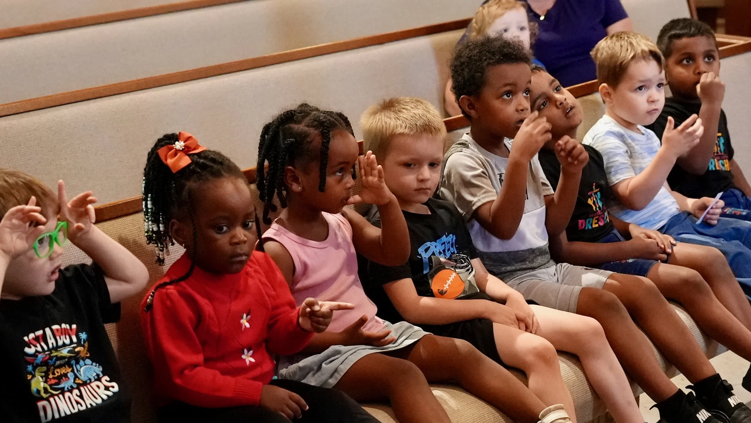 A group of young children at Redeemer Lutheran Preschool eagerly asking questions during a sermon from Pastor Kahle. The children are seated and engaged, listening attentively in a church setting.