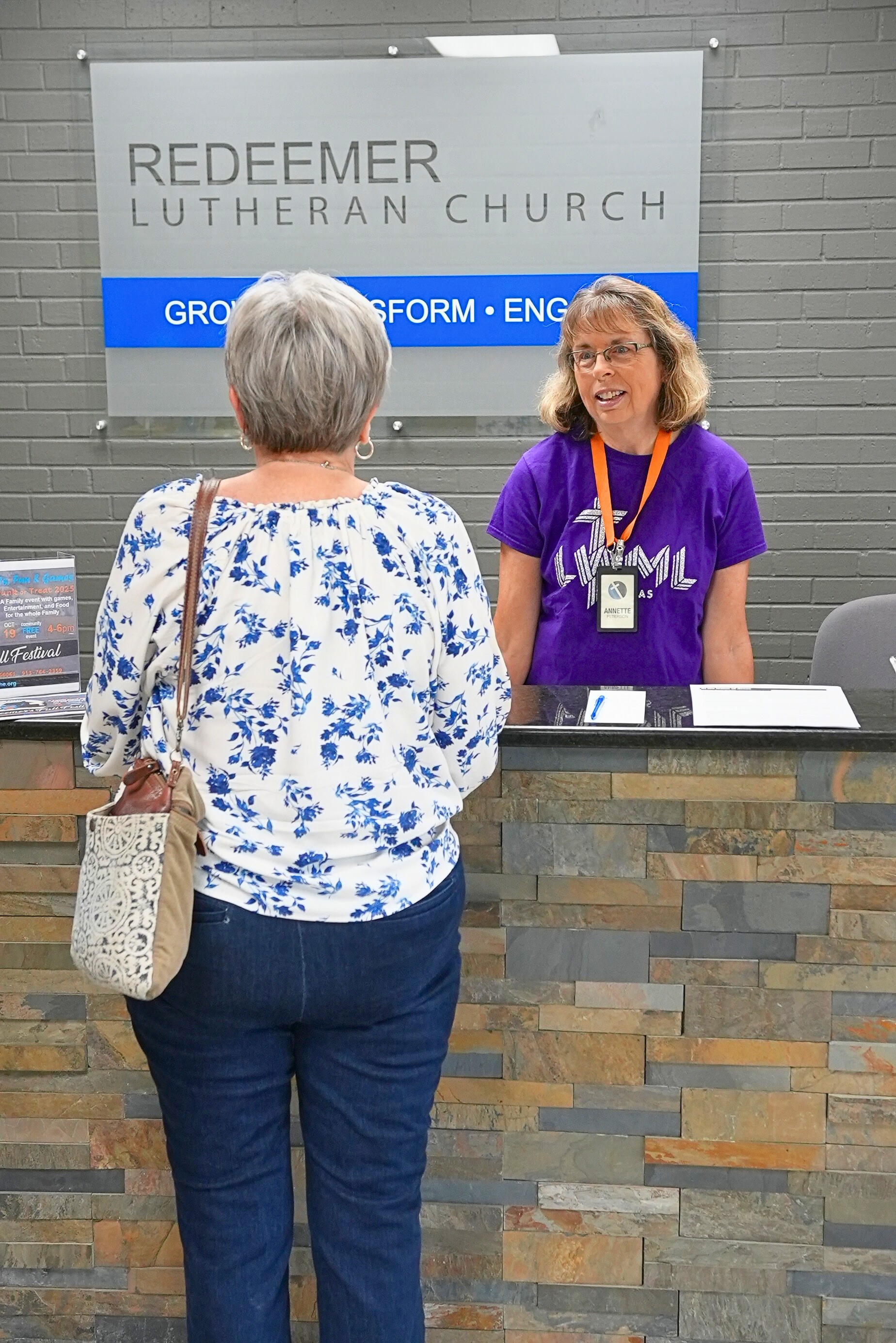 A woman with short gray hair speaking with a staff member at the reception desk inside Redeemer Lutheran Church in Olathe, Kansas. The church environment is welcoming, with bright lighting and a friendly atmosphere for visitors.