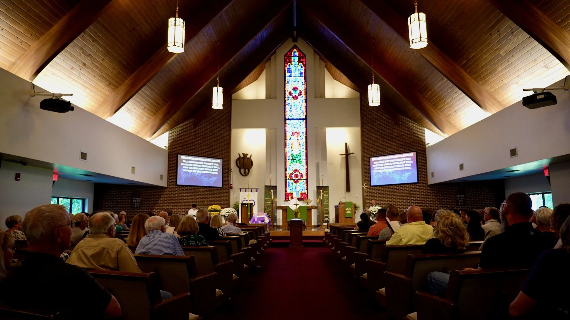 Interior of Redeemer Lutheran Church in Olathe, Kansas, showing wooden ceiling beams, a stained glass cross at the front, and congregation members seated in pews during a service.