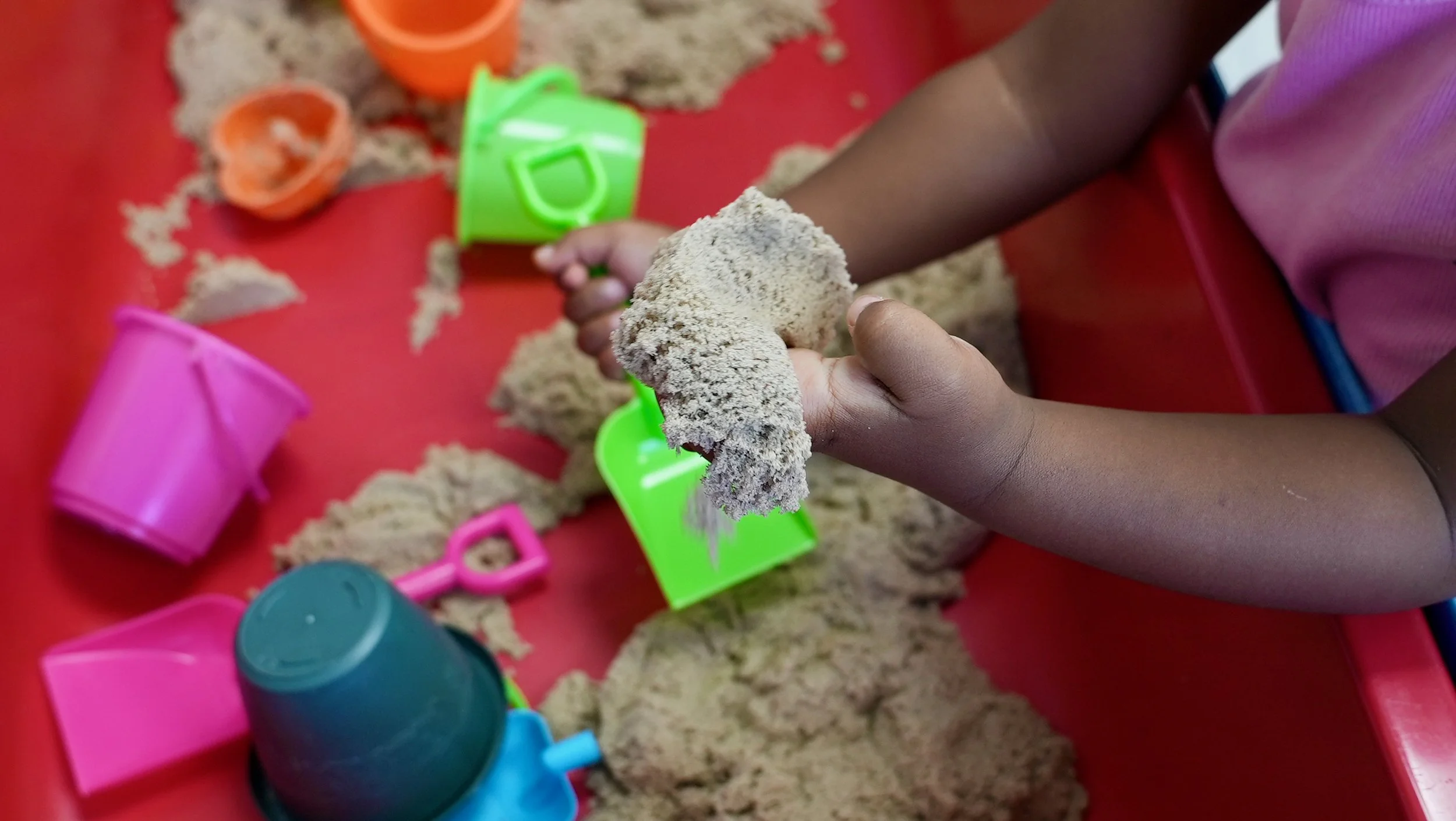 Child playing with sand and plastic toy buckets and shovels on a red surface.