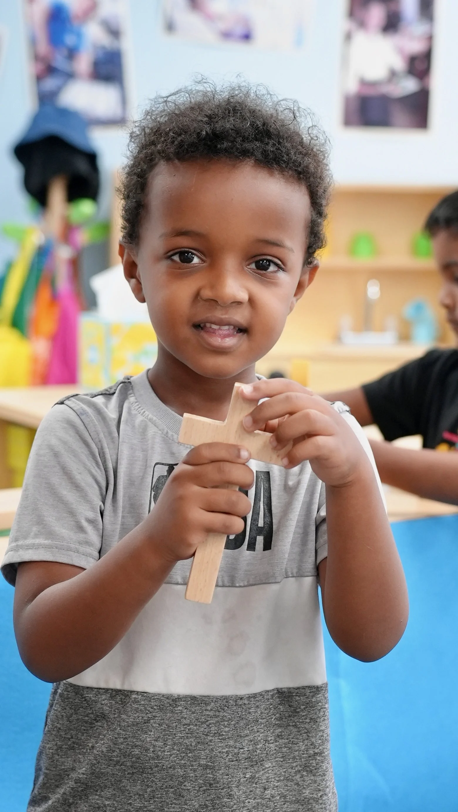 Young boy at Redeemer Lutheran Preschool in Olathe, Kansas, with curly hair and a gray t-shirt holding a wooden cross in a classroom setting.