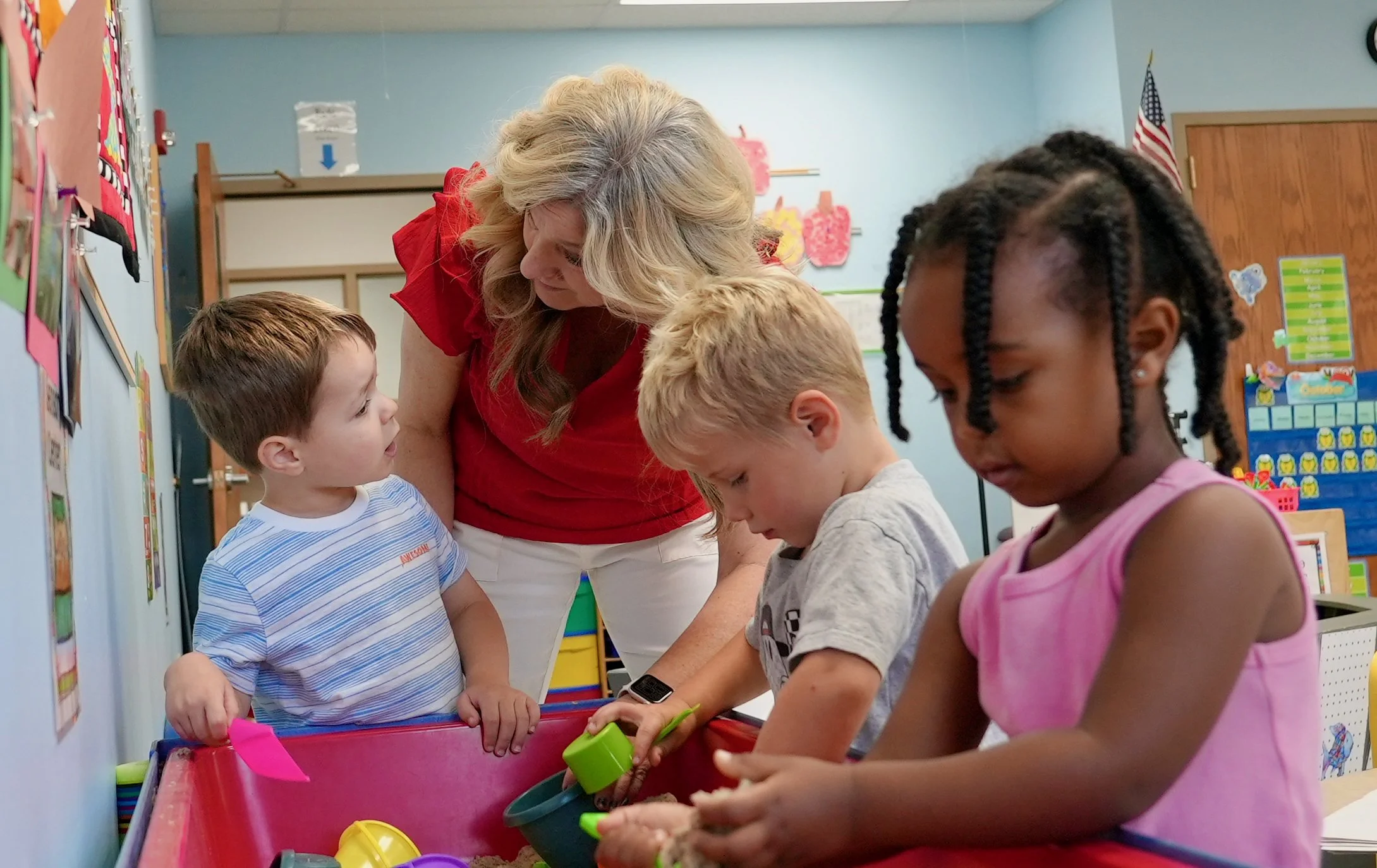 A teacher with three young students in a classroom, engaging in a sensory activity with sand and toys.
