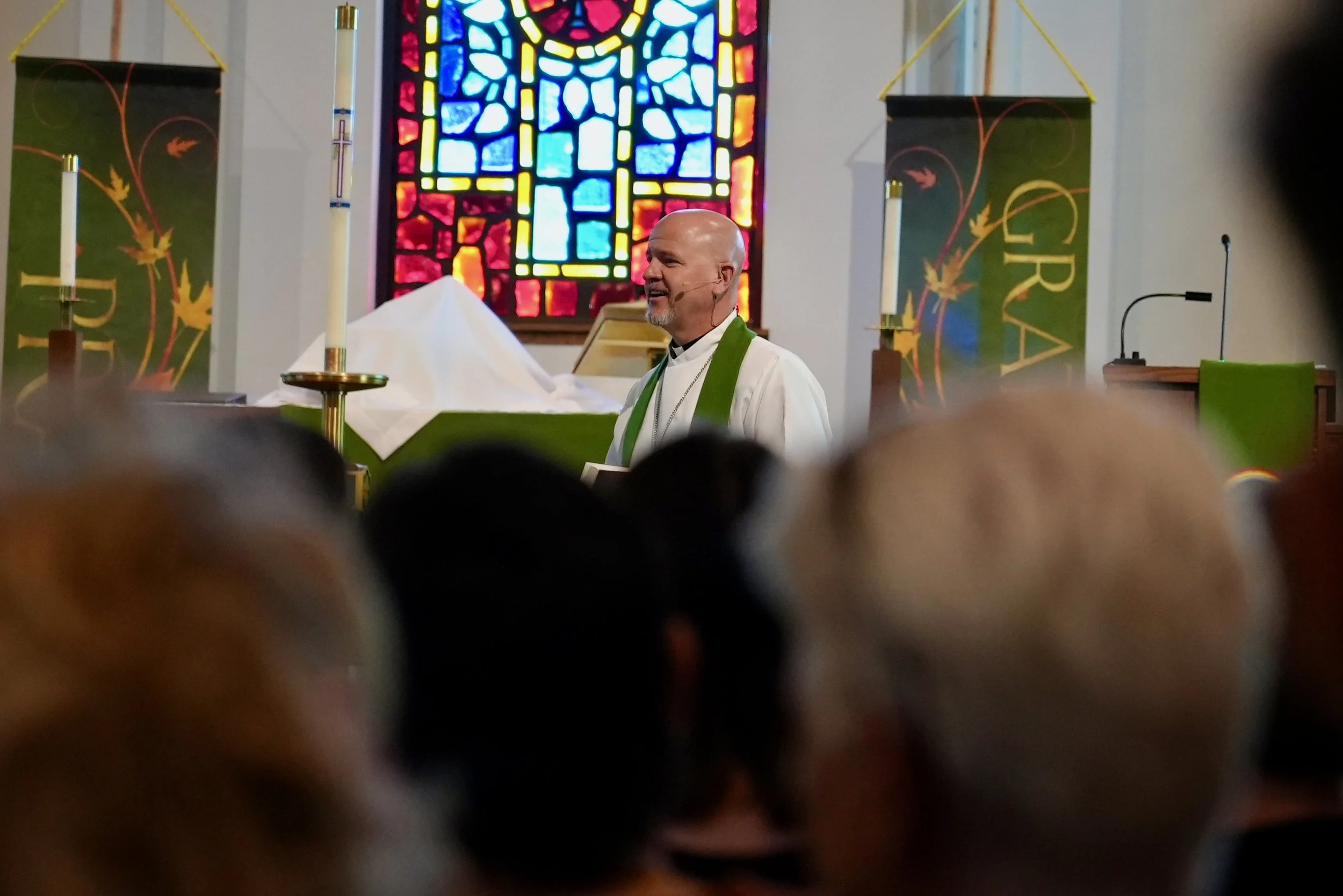 A priest standing at the altar inside a church with a colorful stained glass window in the background, during a service, with an audience in the foreground.