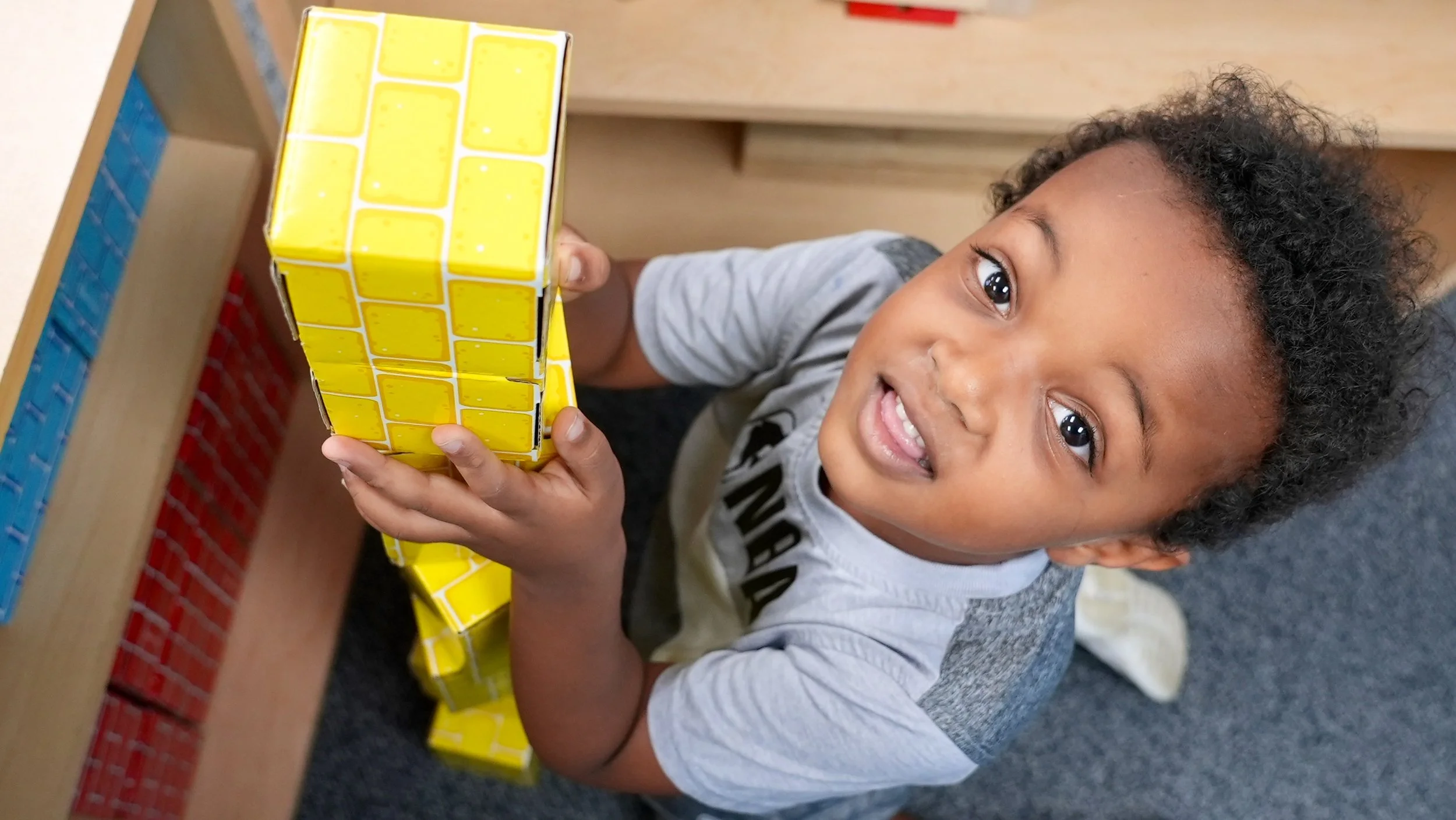 A happy young boy at Redeemer Lutheran Preschool playing with a yellow Rubik’s Cube puzzle. He is smiling, kneeling on the classroom floor near a wooden table, enjoying hands-on learning and play.