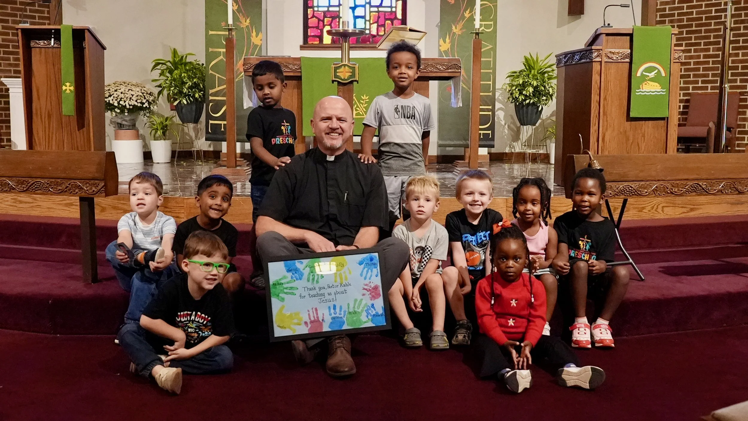 Pastor Kahle with children at Redeemer Lutheran Church, engaging with the kids on stage during a special church activity while the choir director holds a thank-you sign.