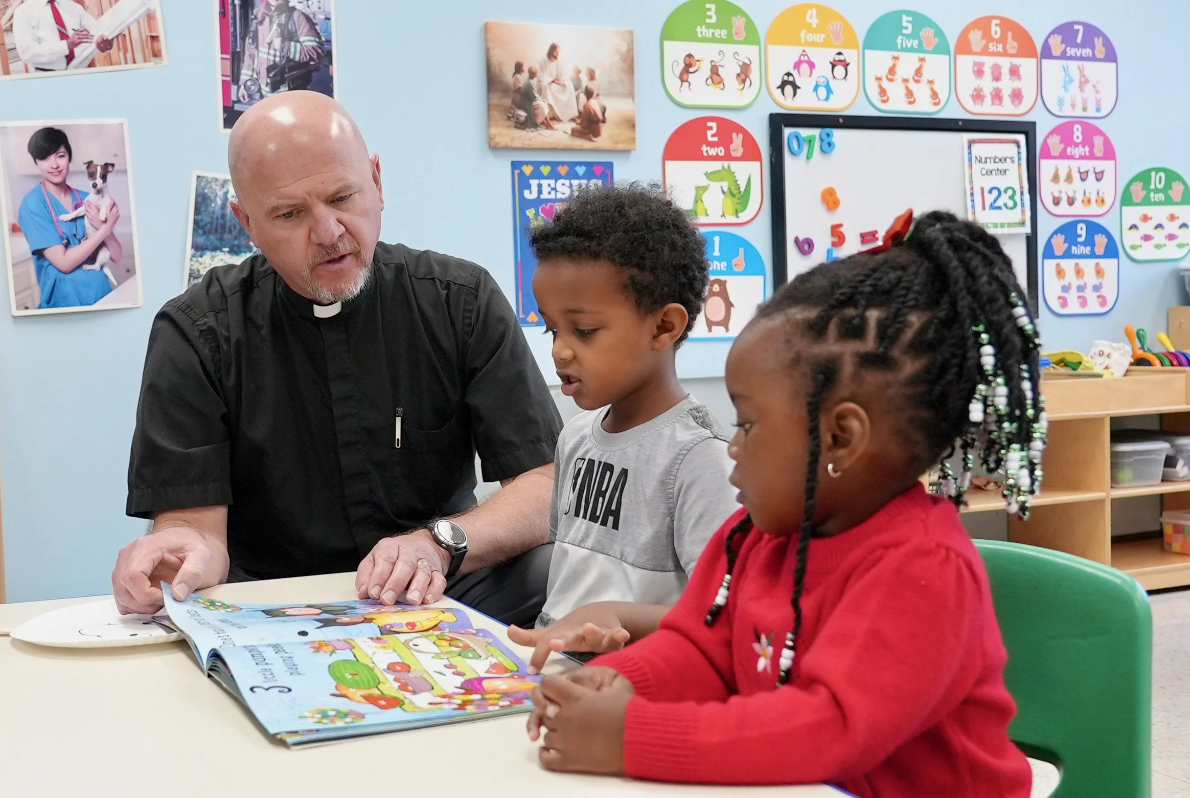 Pastor Kahle at Redeemer Lutheran Preschool engaging with young students, reading a colorful book together at a classroom table. The classroom features educational posters, numbers, and animal illustrations, supporting early childhood learning and fa