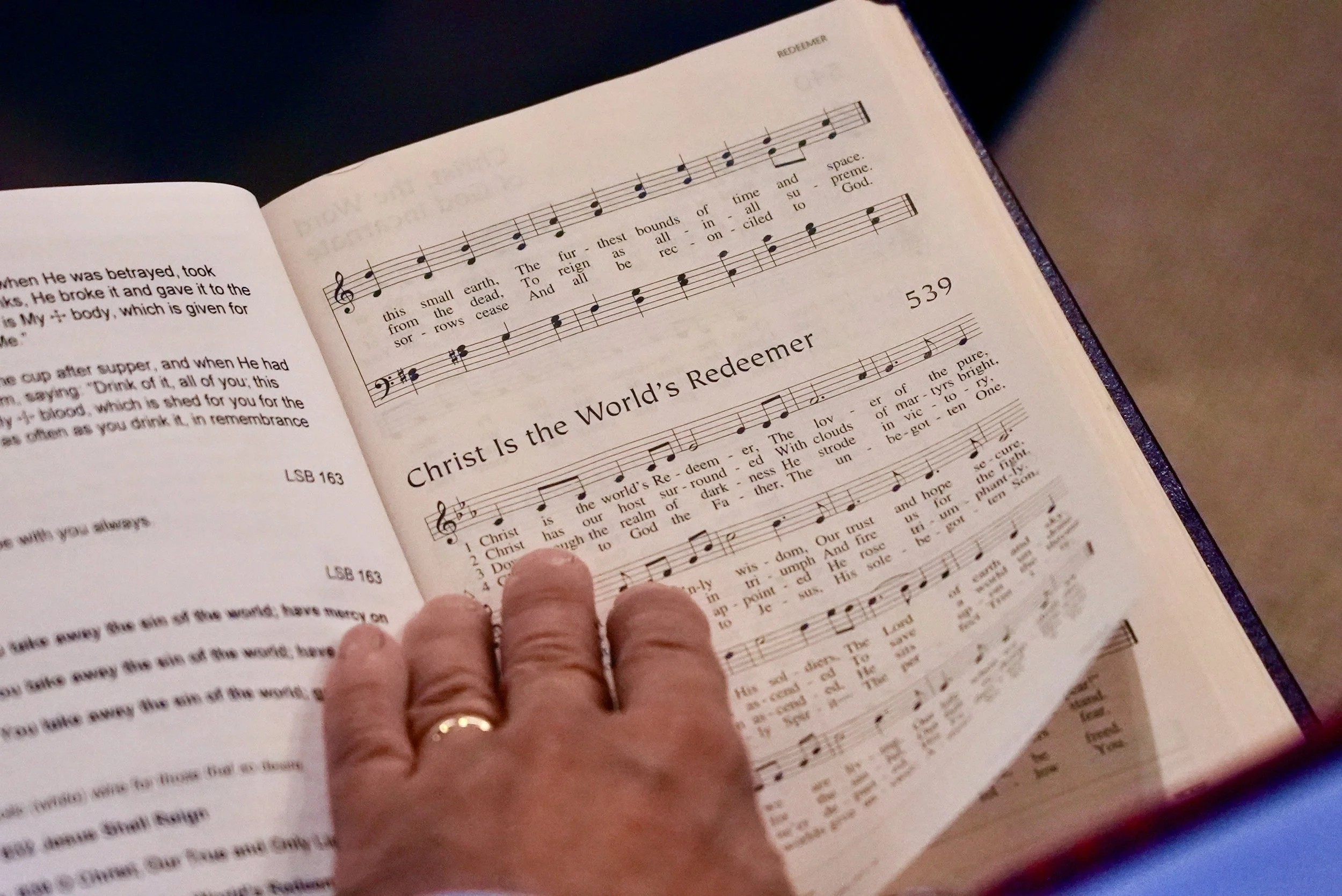 A person with a wedding ring turns a page in a hymnal or songbook, displaying the sheet music for 'Christ Is the World's Redeemer' at Redeemer Lutheran Church in Olathe, Kansas.