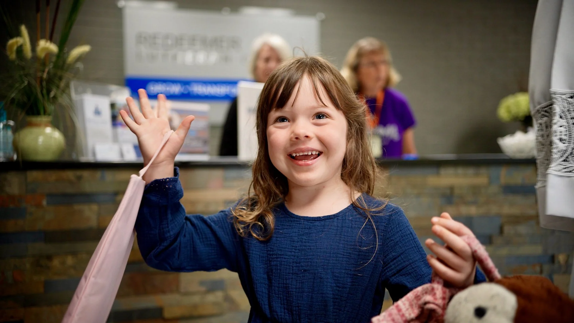A young girl entering Redeemer Lutheran Church, smiling and waving at the reception desk, with two staff members visible behind the counter.