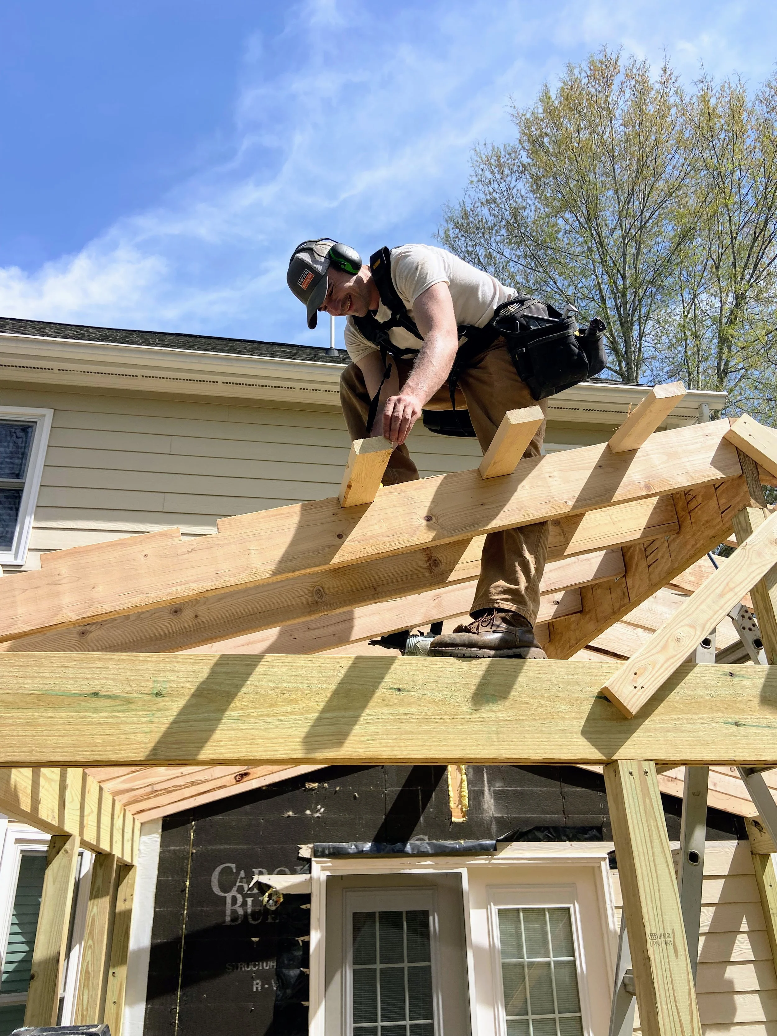 A man wearing a helmet, headphones, and work gear is working on the wooden framework of a roof of a house under construction, with a blue sky and trees in the background.