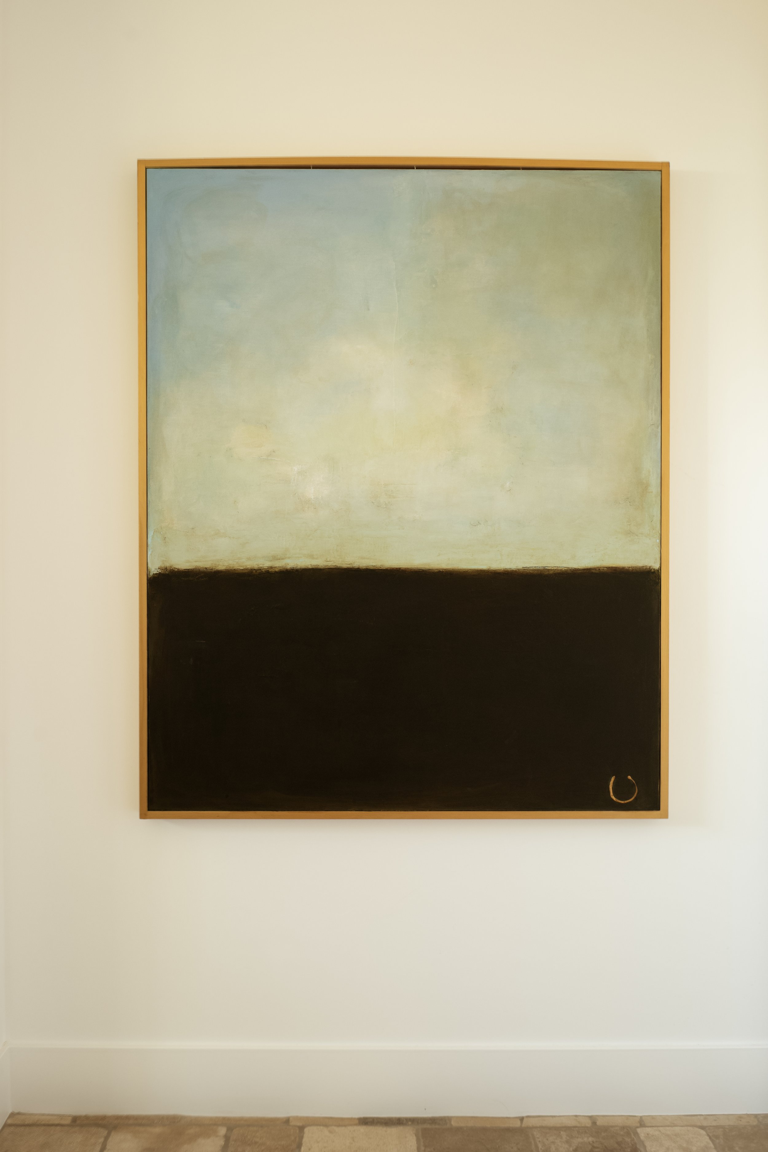 Abstract painting on a wall with a large light blue-green section above a dark black-brown section, framed in wood, with a small gold circle in the bottom right corner.