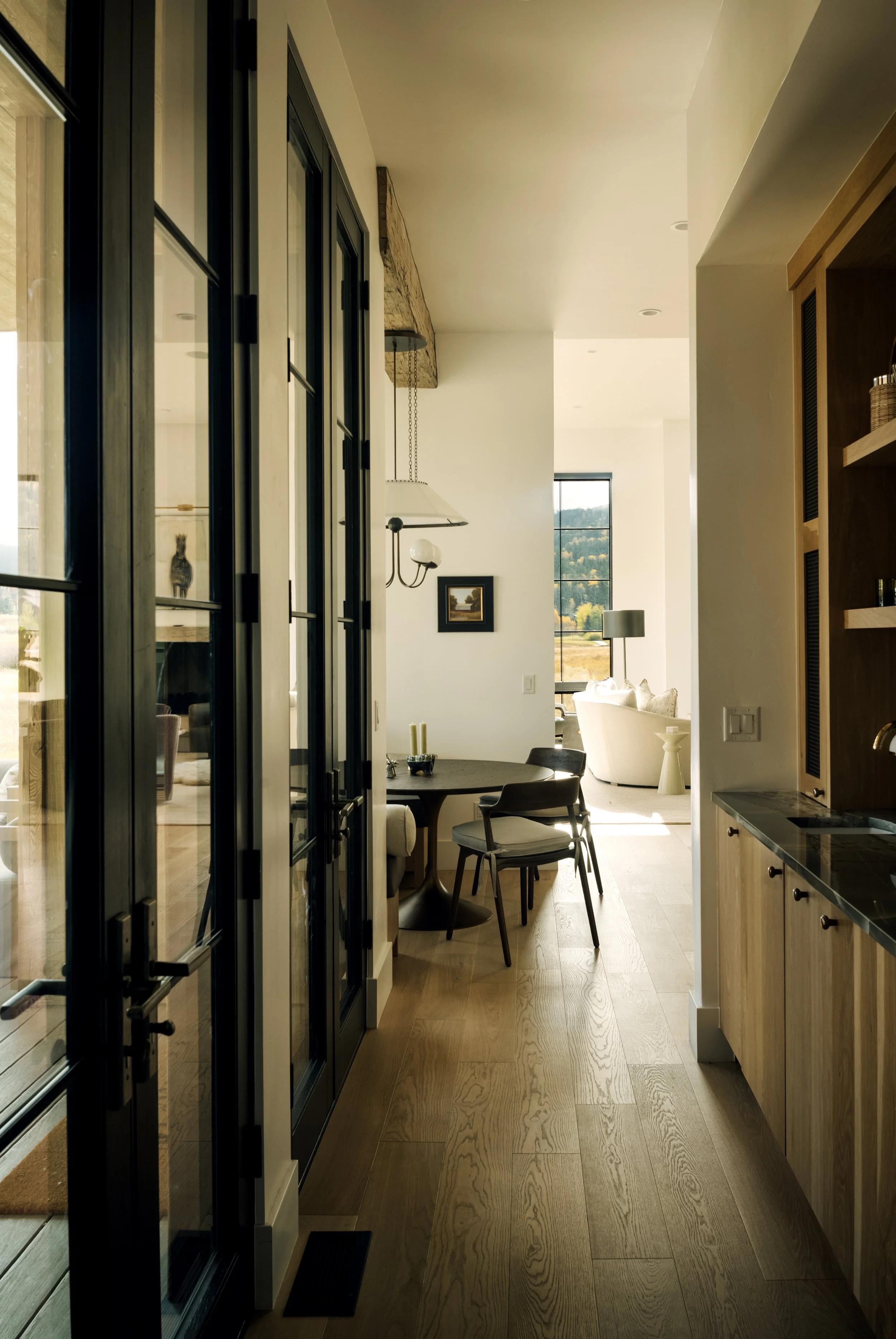 Interior view of a modern home with a wooden floor, black-framed glass doors, a dining area with a round table and chairs, and a living room with a white sofa and large window showing a scenic landscape.