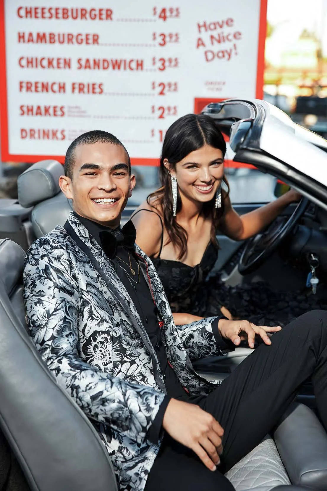 A young man and woman sitting in a convertible car, dressed in formal attire, smiling at the camera. Behind them is a red and white menu board with food prices and the message 'Have a Nice Day!'
