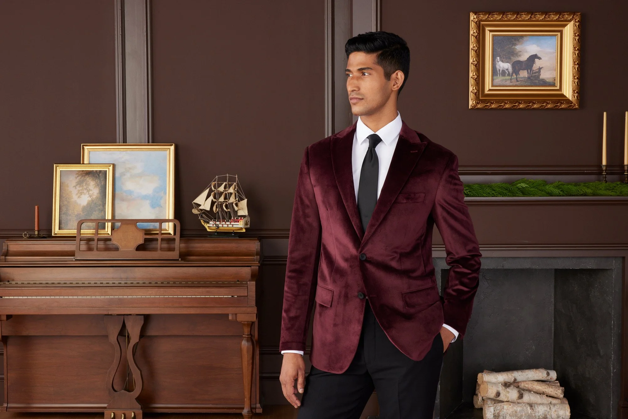 A man wearing a burgundy velvet blazer, white shirt, black tie, and black pants, standing in front of a wooden piano and framed paintings on a brown wall, with a stack of firewood nearby.