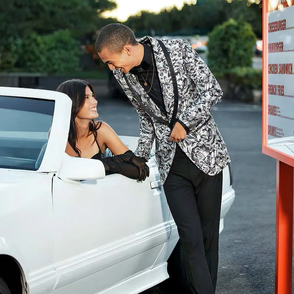A woman sitting in a white convertible car, smiling and looking up at a man standing next to her. The man is leaning slightly towards her, smiling back, wearing a floral patterned jacket. They are outdoors near a parking lot with greenery in the back