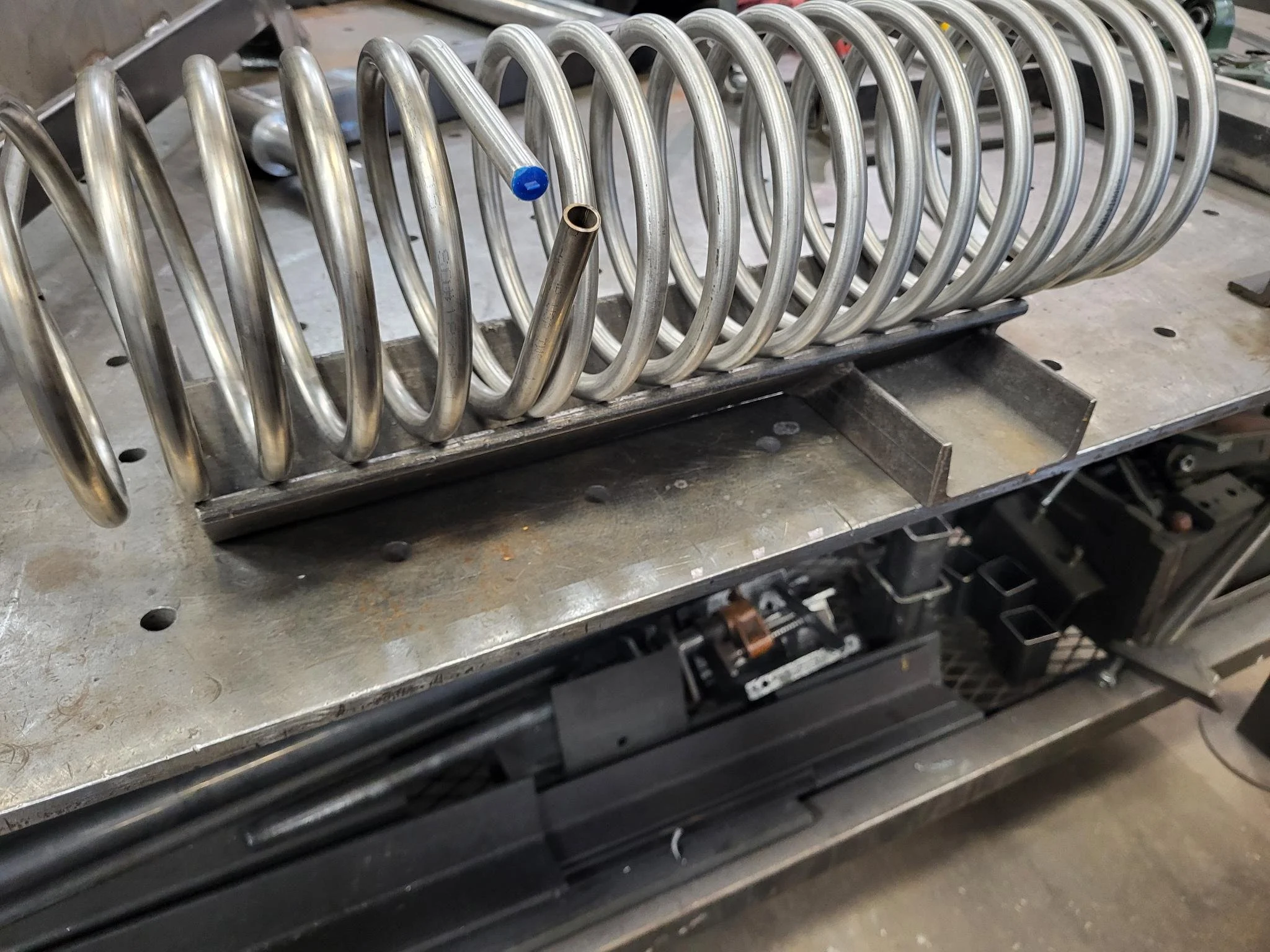 Metalwork coil with a blue cap and tubing, mounted on a metal table in what appears to be a workshop or manufacturing setting.