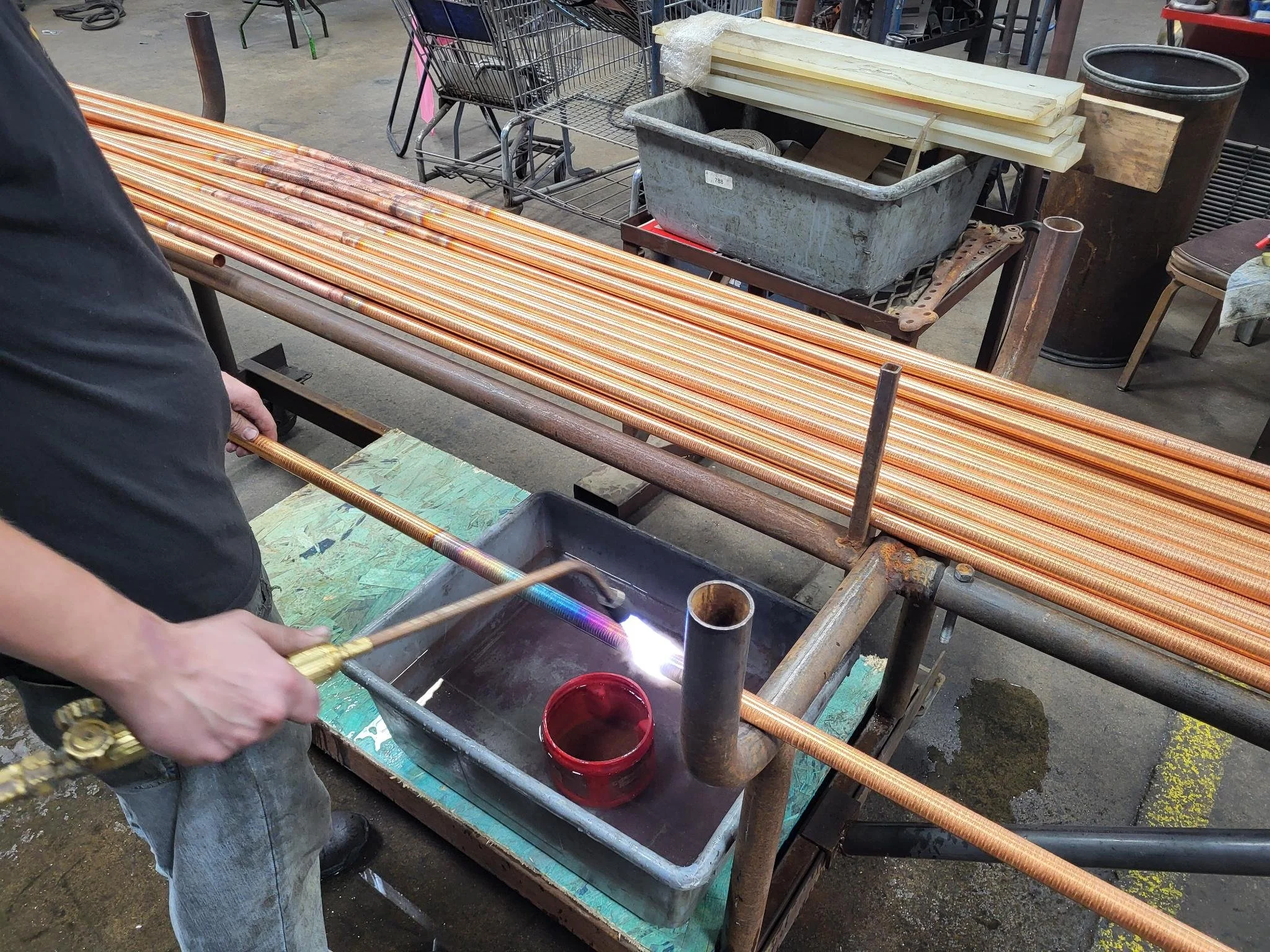 A person welding copper pipes inline in a workshop, with coiled copper pipes on the table and various tools and materials around.