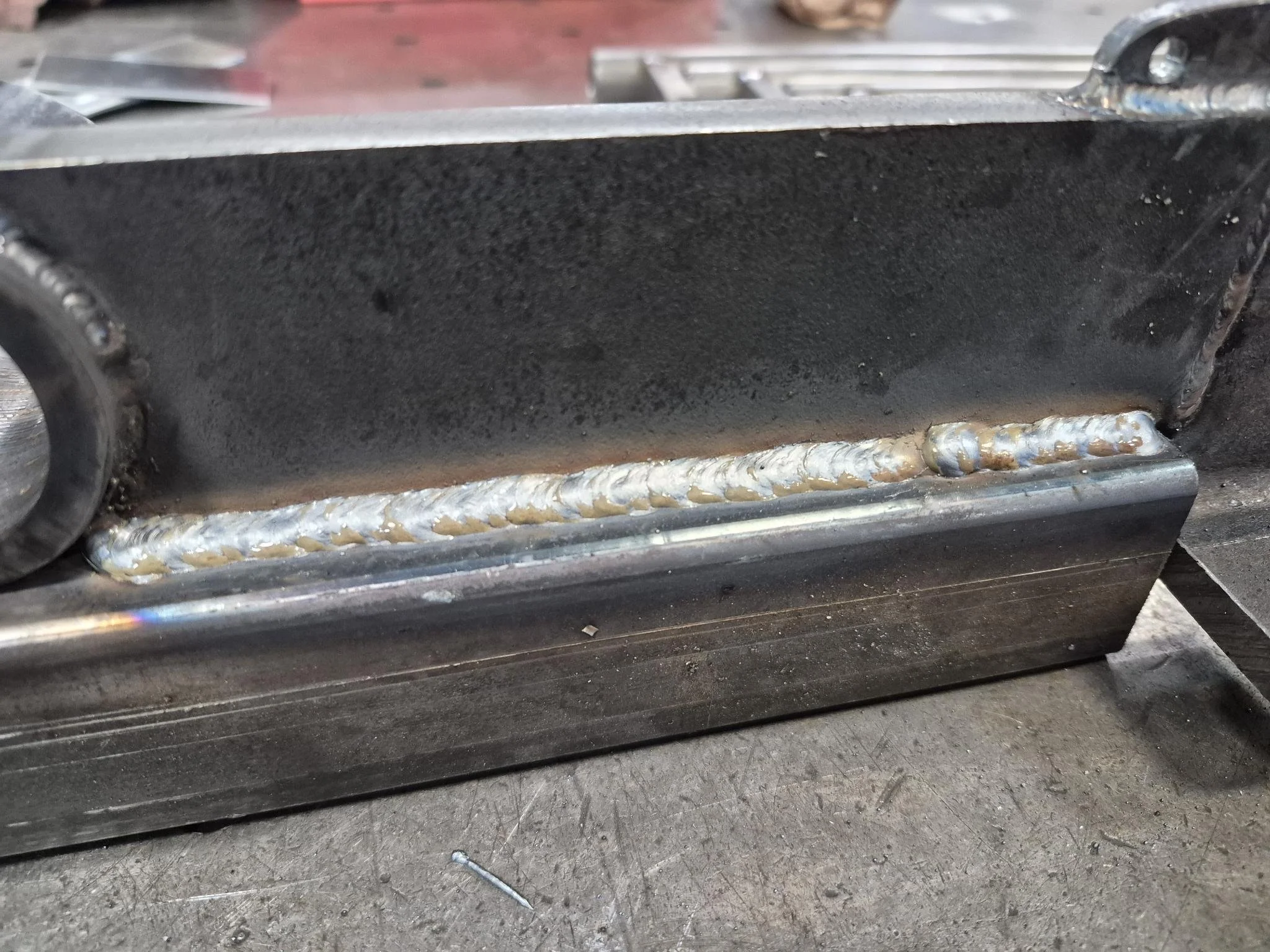 Close-up of a metal component with a long weld bead along the edge, showing a rough, metallic surface and some discoloration, in a workshop setting.