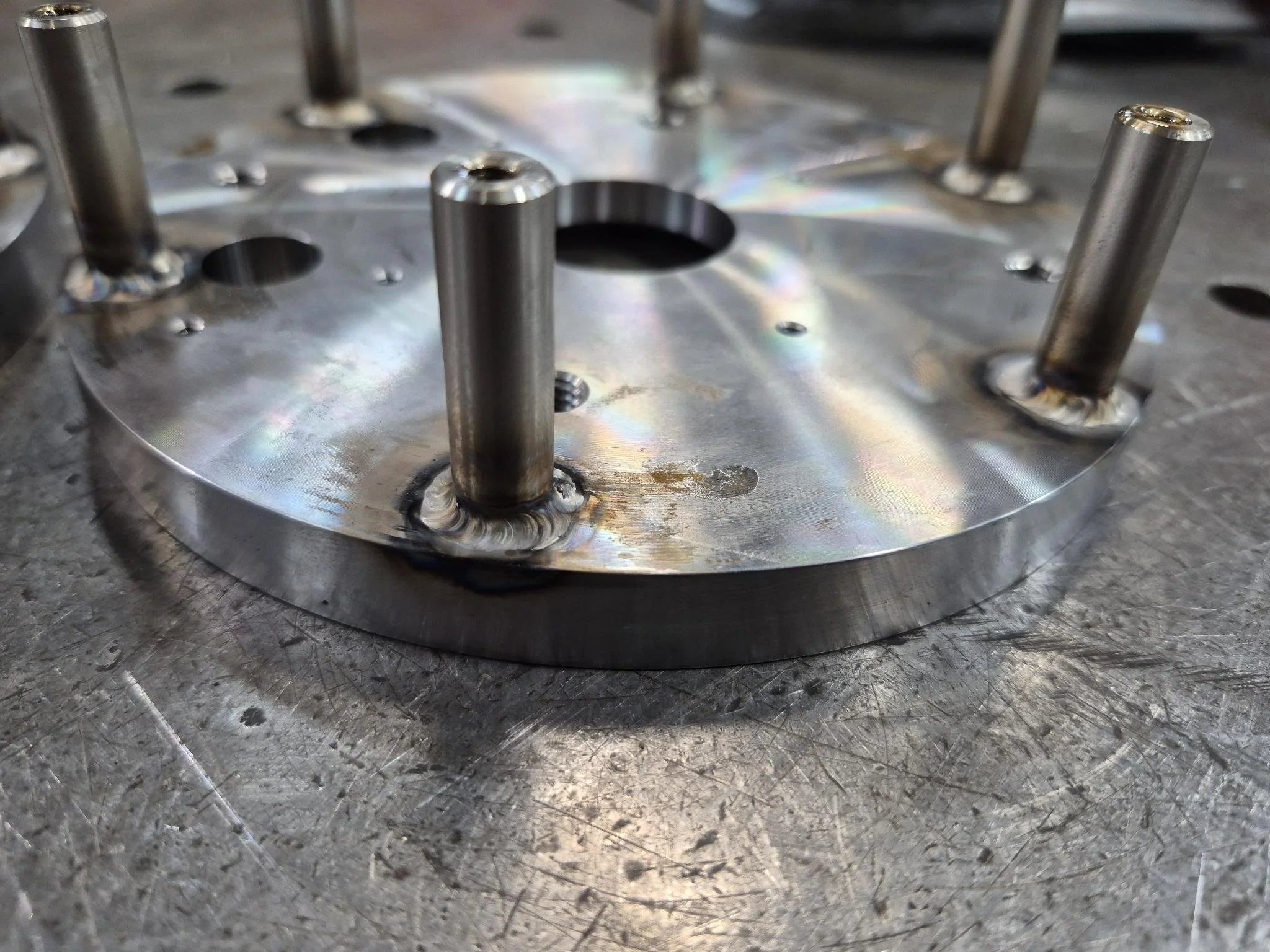 Close-up of a metal mechanical component with threaded rods welded onto a flat metal surface. The surface has a rainbow sheen from the welding process, and the background shows a textured metal worktable.