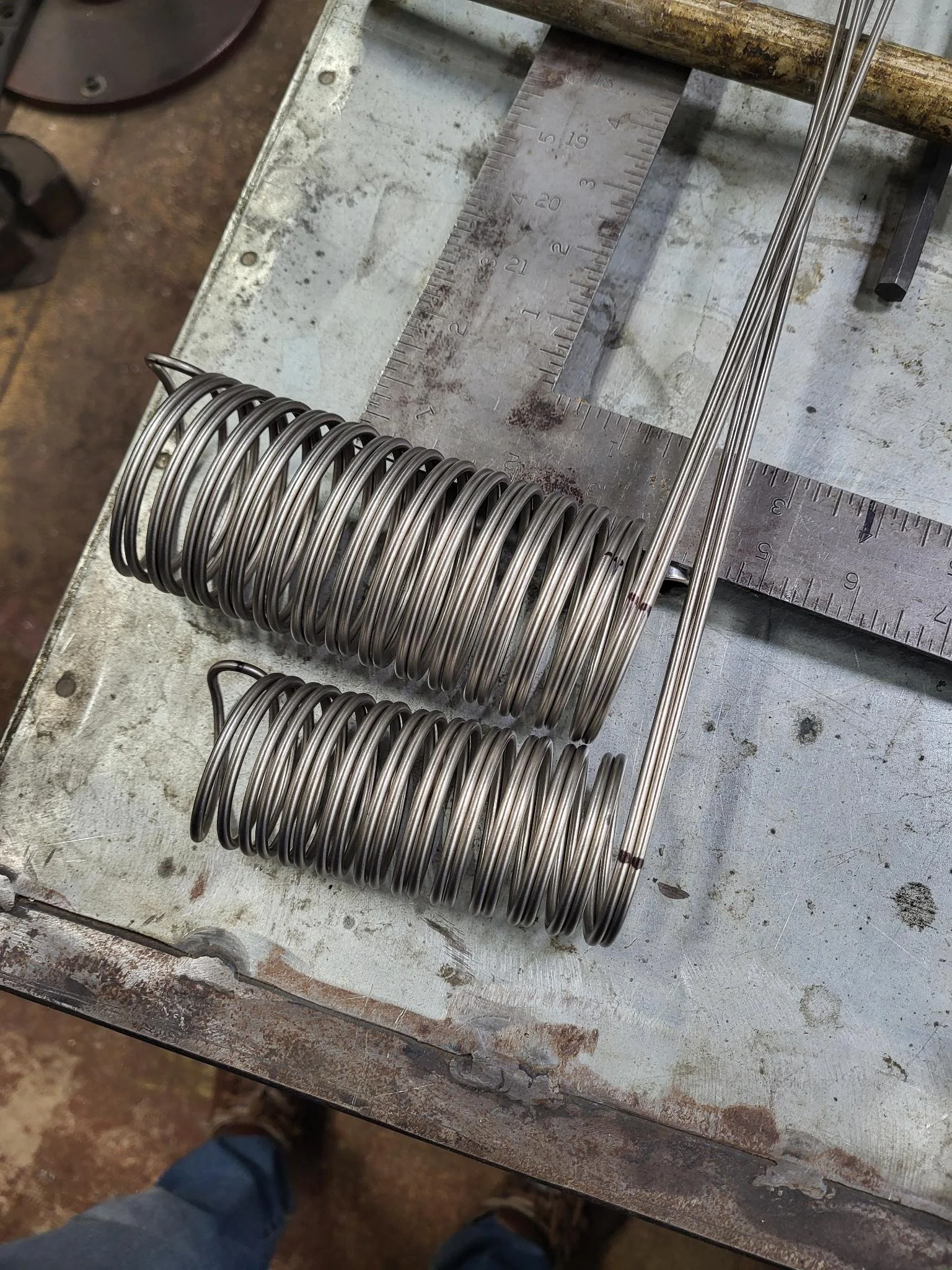 Two metal coil springs and two metal rods on a worktable with a metal square and ruler for measurement.