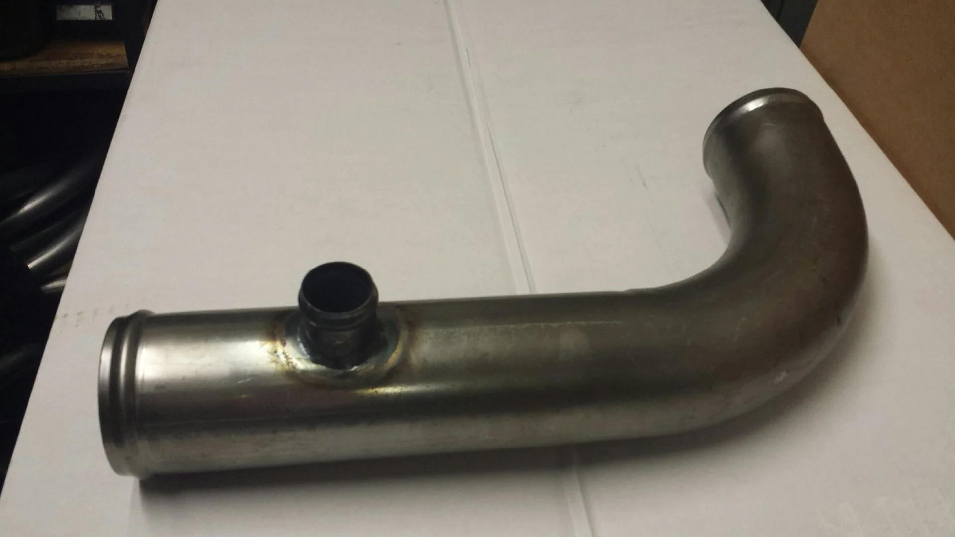 Metal pipe with a bend and a small opening on one side, lying on a white surface.