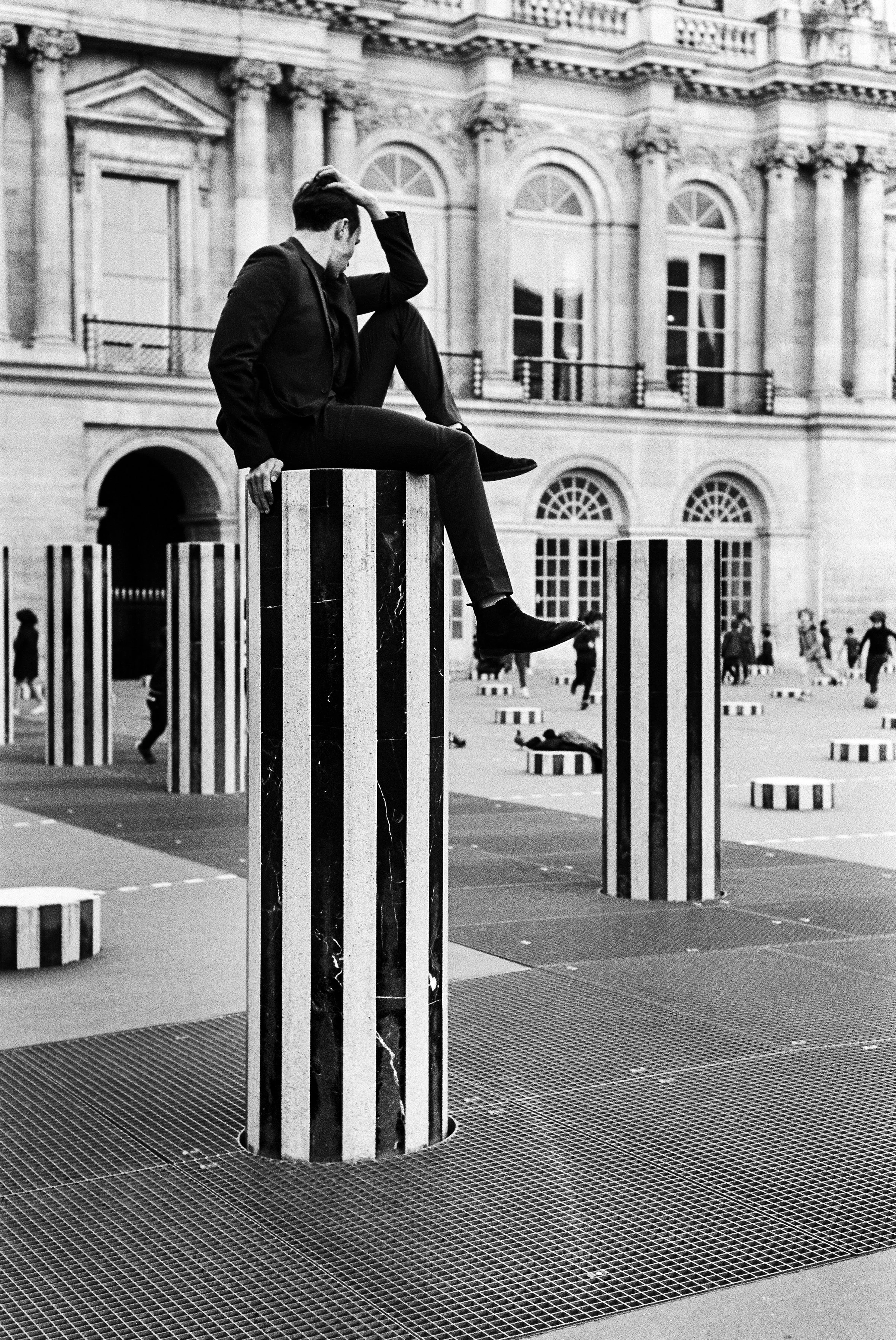 A man dressed in a suit is sitting on a striped black and white column in an outdoor plaza with historical buildings in the background. The plaza features multiple similar striped columns and circular platforms, with people walking and relaxing around.
