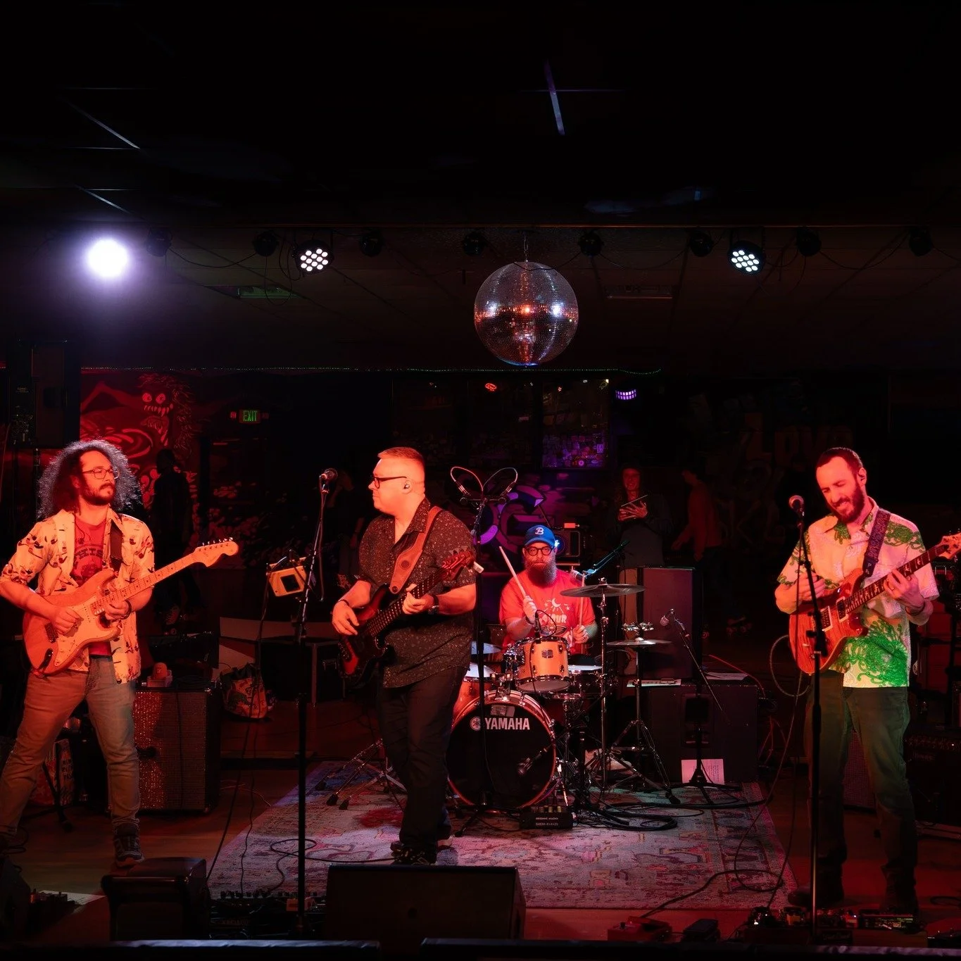 A four-piece band performing live on stage with guitars and drums, under a disco ball and stage lights in a dimly lit venue
