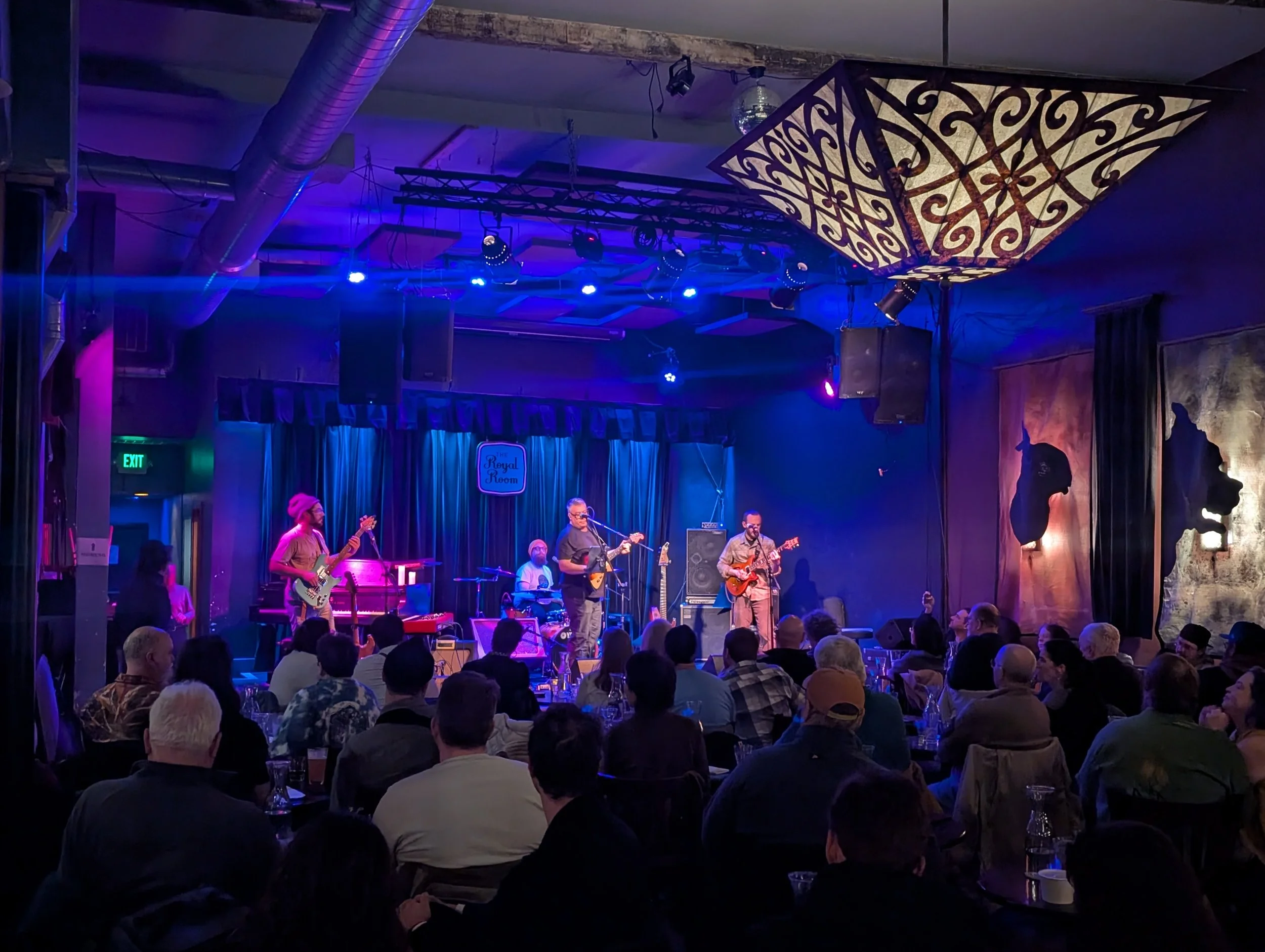 A live musical performance on stage with four musicians playing instruments, including two guitarists, a bassist, and a keyboardist, in front of an audience in a dimly lit venue with blue and purple lighting, curtains, and artwork on the walls.