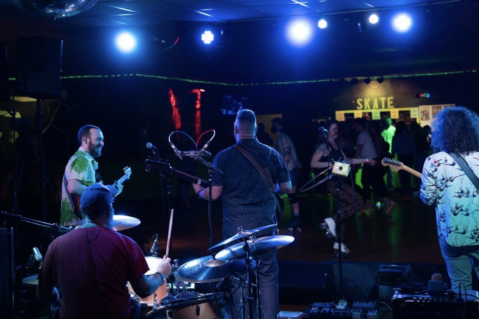 Musicians performing live on stage at a nightclub, with people dancing in the background and a sign that says 'SKATE' in the room.