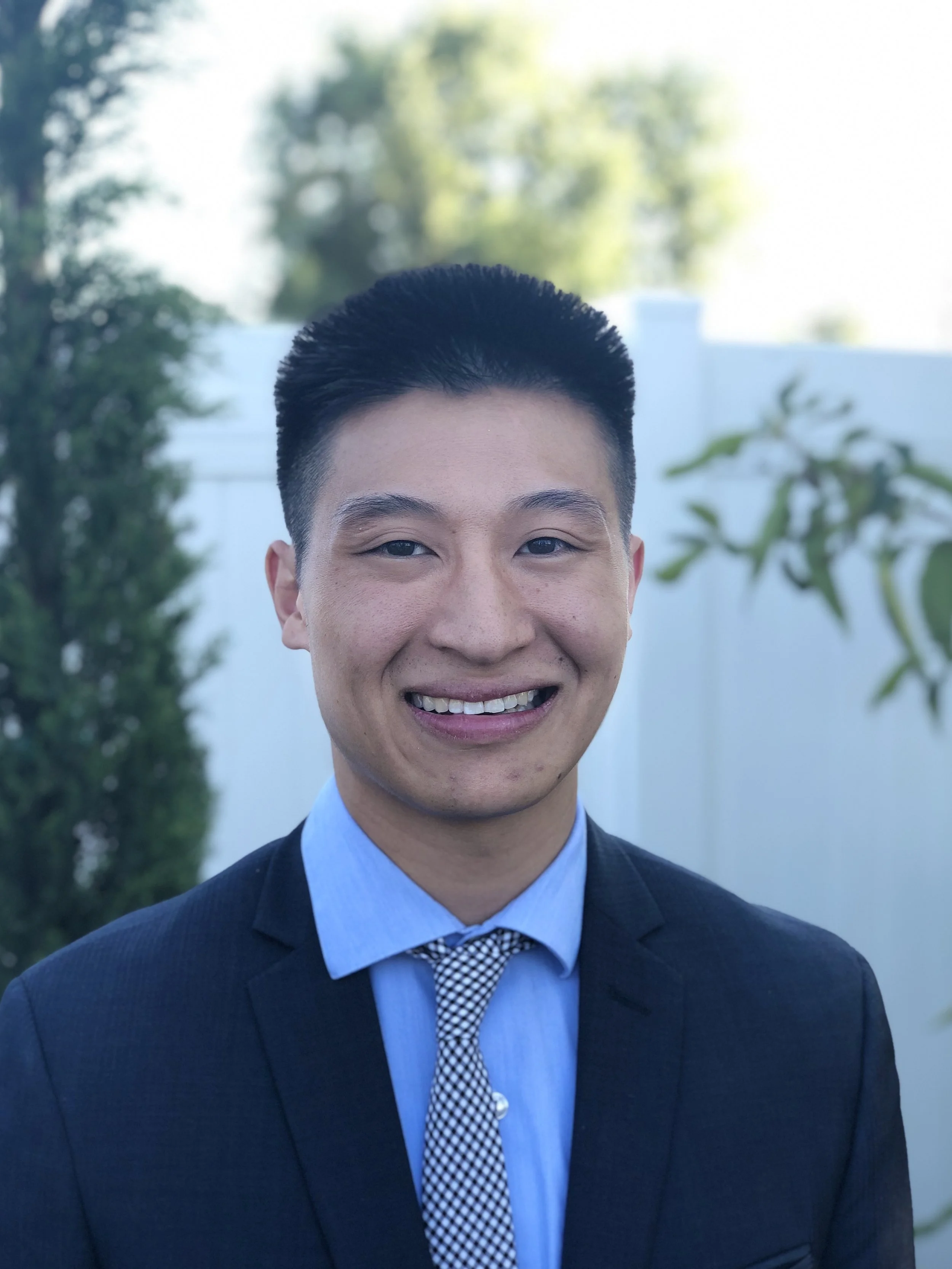 A young Asian man in a dark suit, light blue shirt, and checkered tie smiling outdoors with greenery and a white fence in the background.