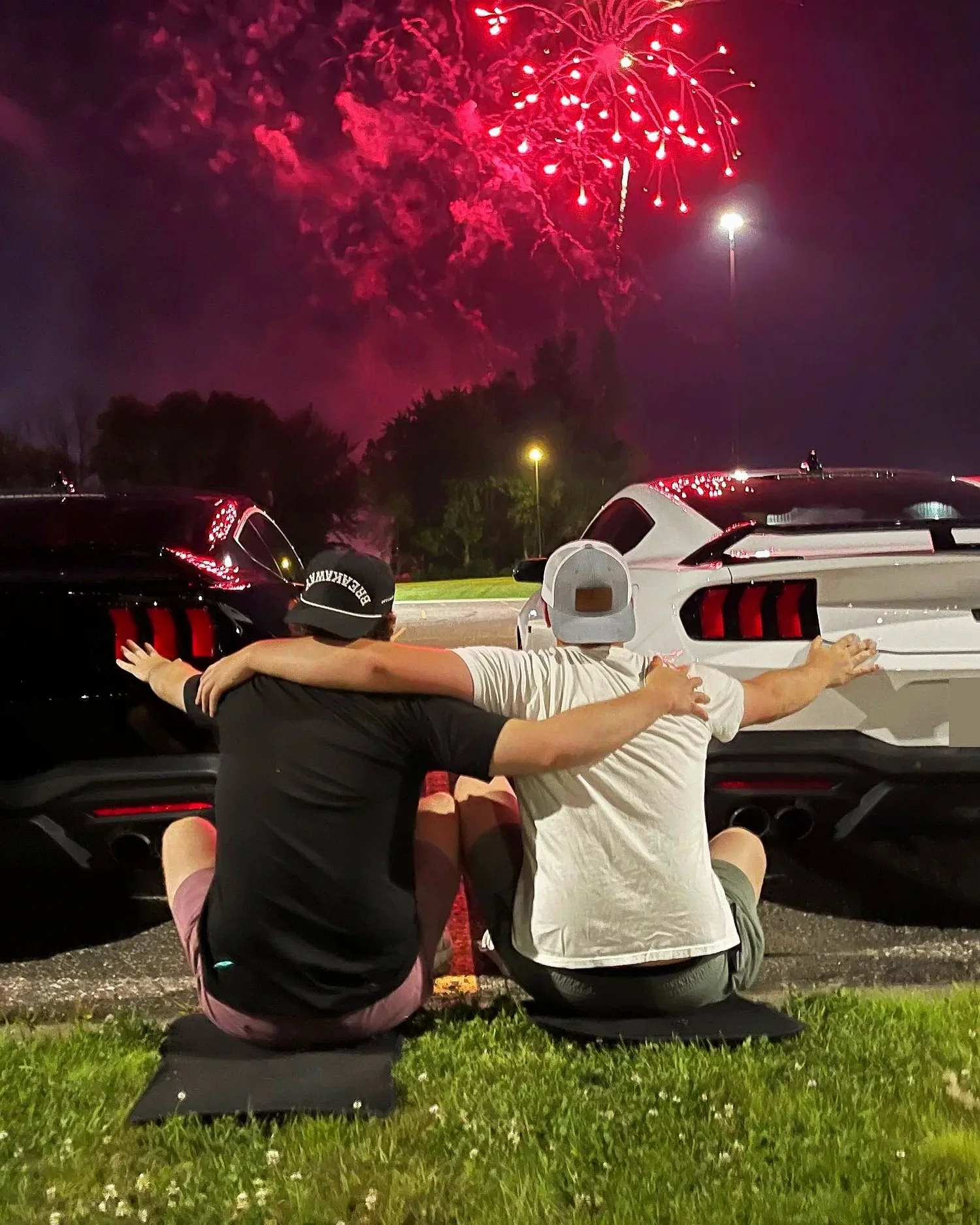Two people sitting on the ground with their arms around each other, watching fireworks in the night sky near two parked cars.