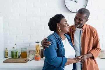 An older couple smiling and holding hands in a kitchen with white brick walls, jars of spices, and cooking ingredients on the counter.