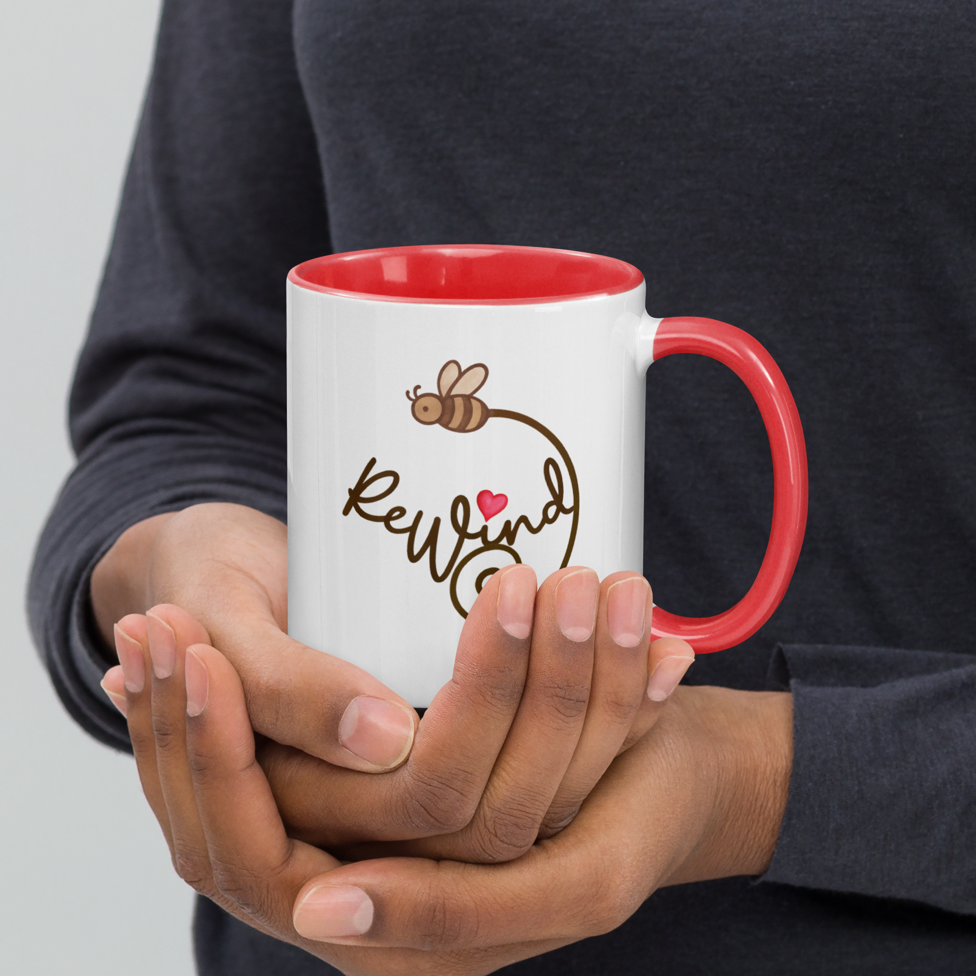 Person holding red-accent mug showing ReWind script with bee trail and heart.