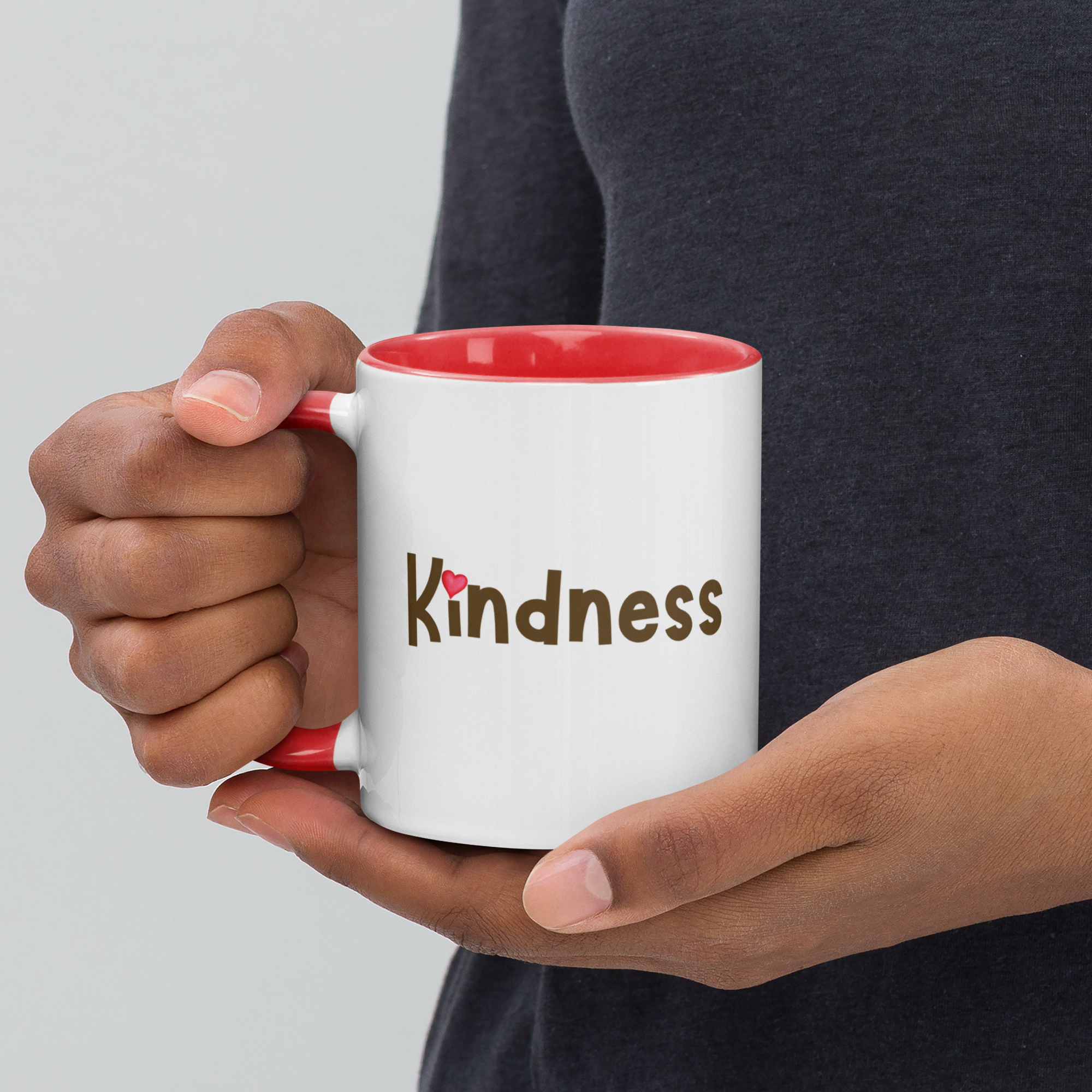 Person holding red-accent mug showing “Kindness” wordmark with heart over the i.