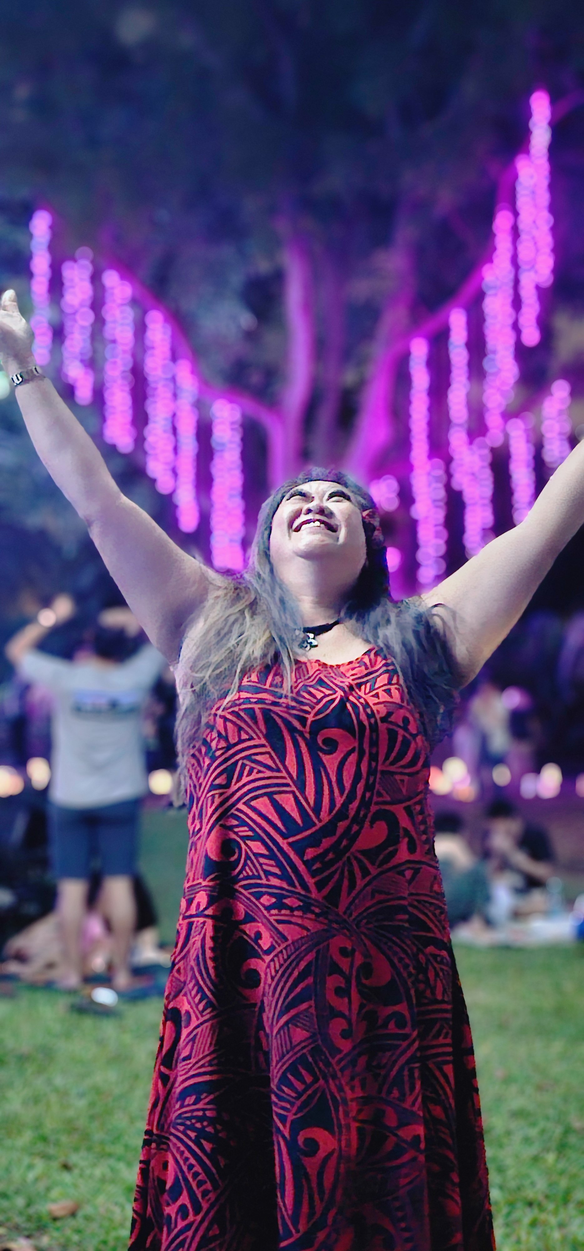 A woman wearing a red and black patterned dress with her arms raised, joyful expression, standing outdoors at a nighttime event with purple lights and trees in the background.