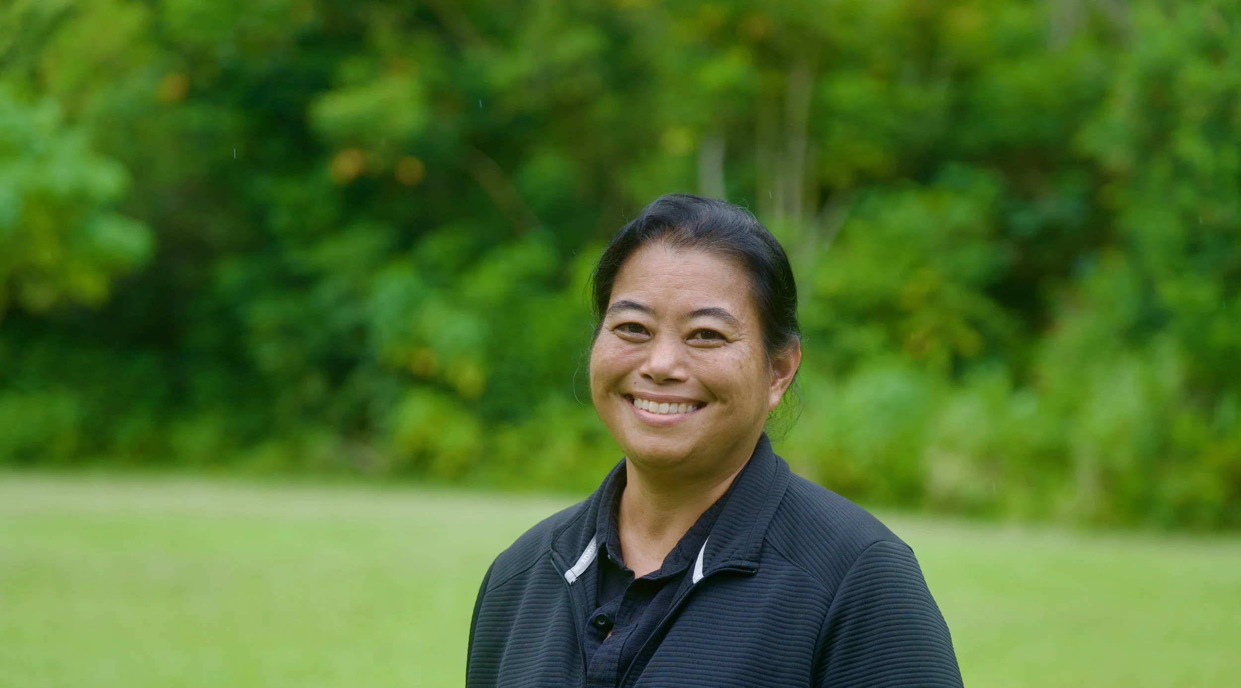 A smiling woman wearing a black jacket outdoors with green trees and grass in the background.