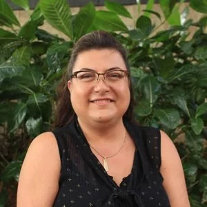 A woman with glasses and dark hair smiling outdoors with green leafy plants in the background.