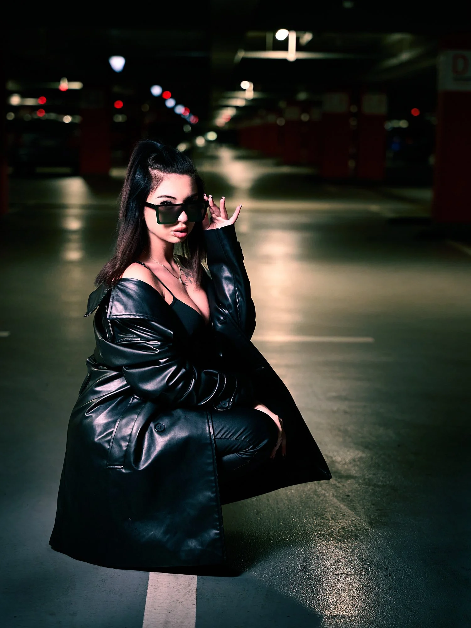 A woman squatting in a parking garage, wearing black sunglasses, a black leather trench coat, and black pants, with dark hair and a confident expression.