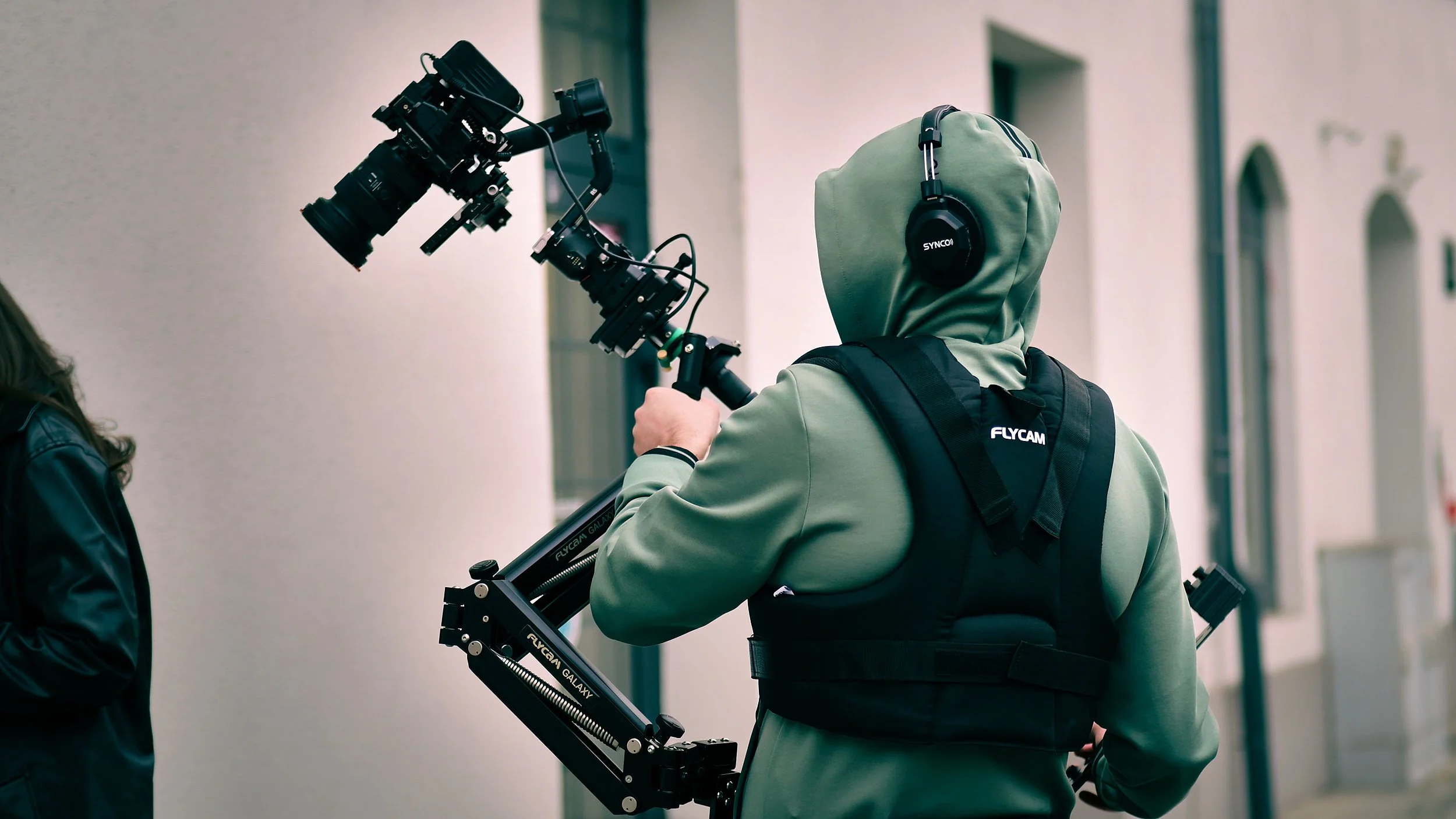 Person wearing a green hoodie, black vest, and headphones operating a camera stabilizer with a mounted camera during a filming session outdoors.