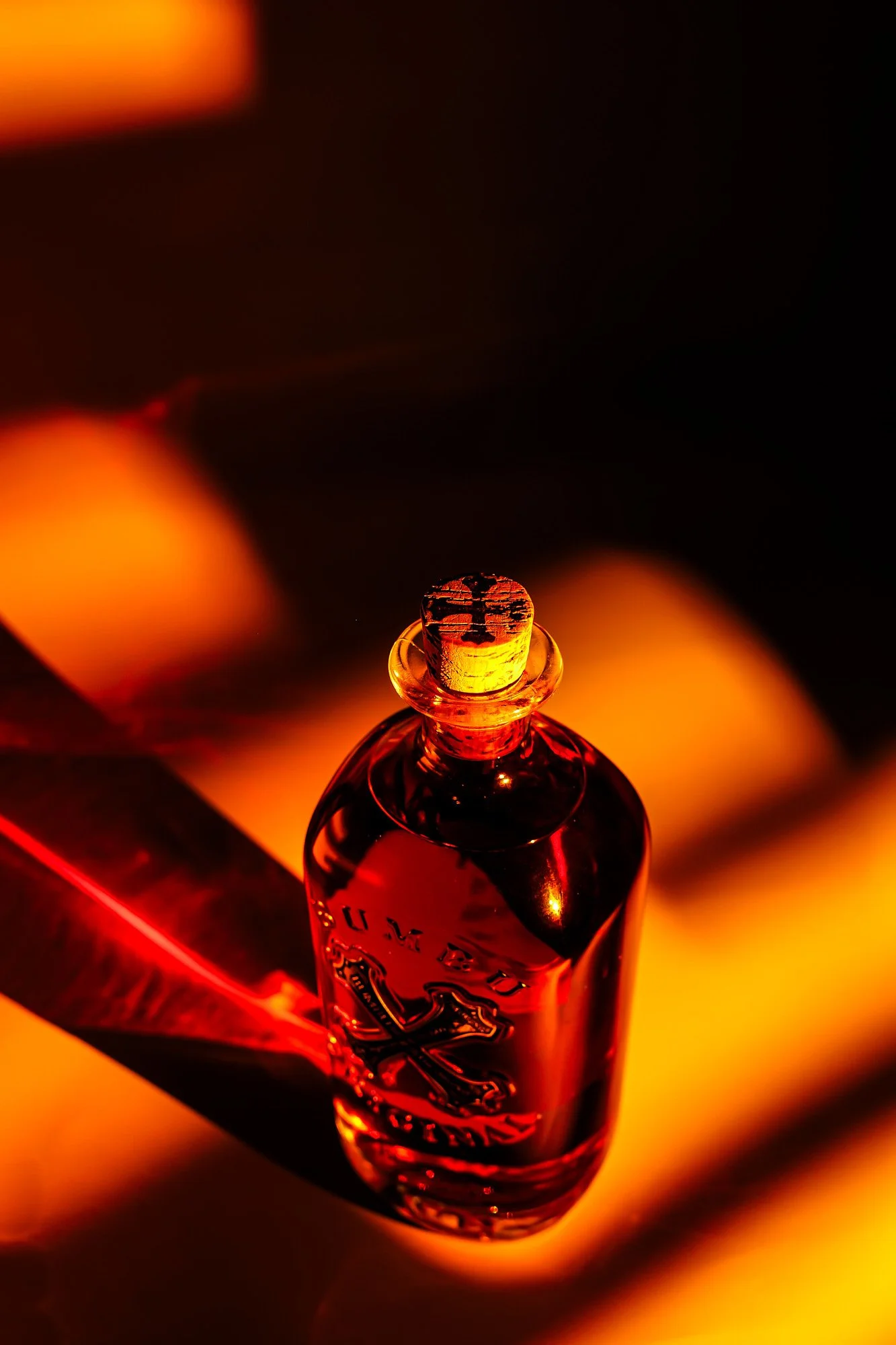 A glass perfume bottle with a cork stopper, illuminated with warm red and orange lighting, casting shadows on the surface.
