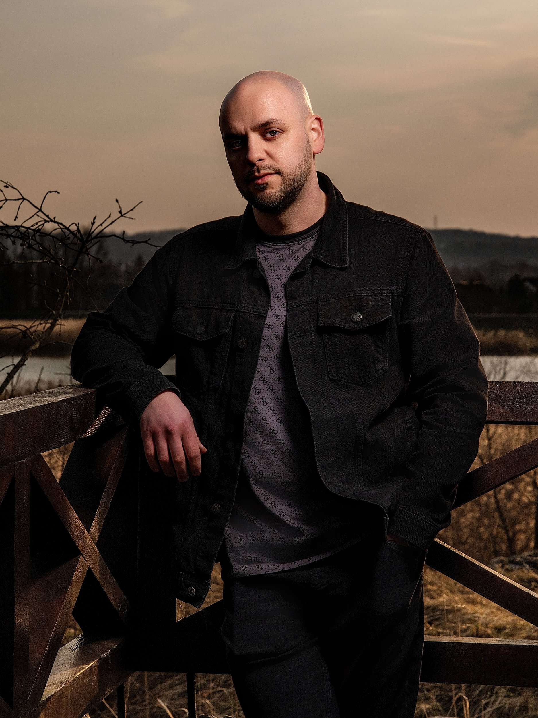 A man with a shaved head and beard stands outdoors at sunset, leaning on a wooden railing. He is wearing a black denim jacket over a patterned gray shirt and has one hand in his pocket. The background features a rural landscape with trees and distant