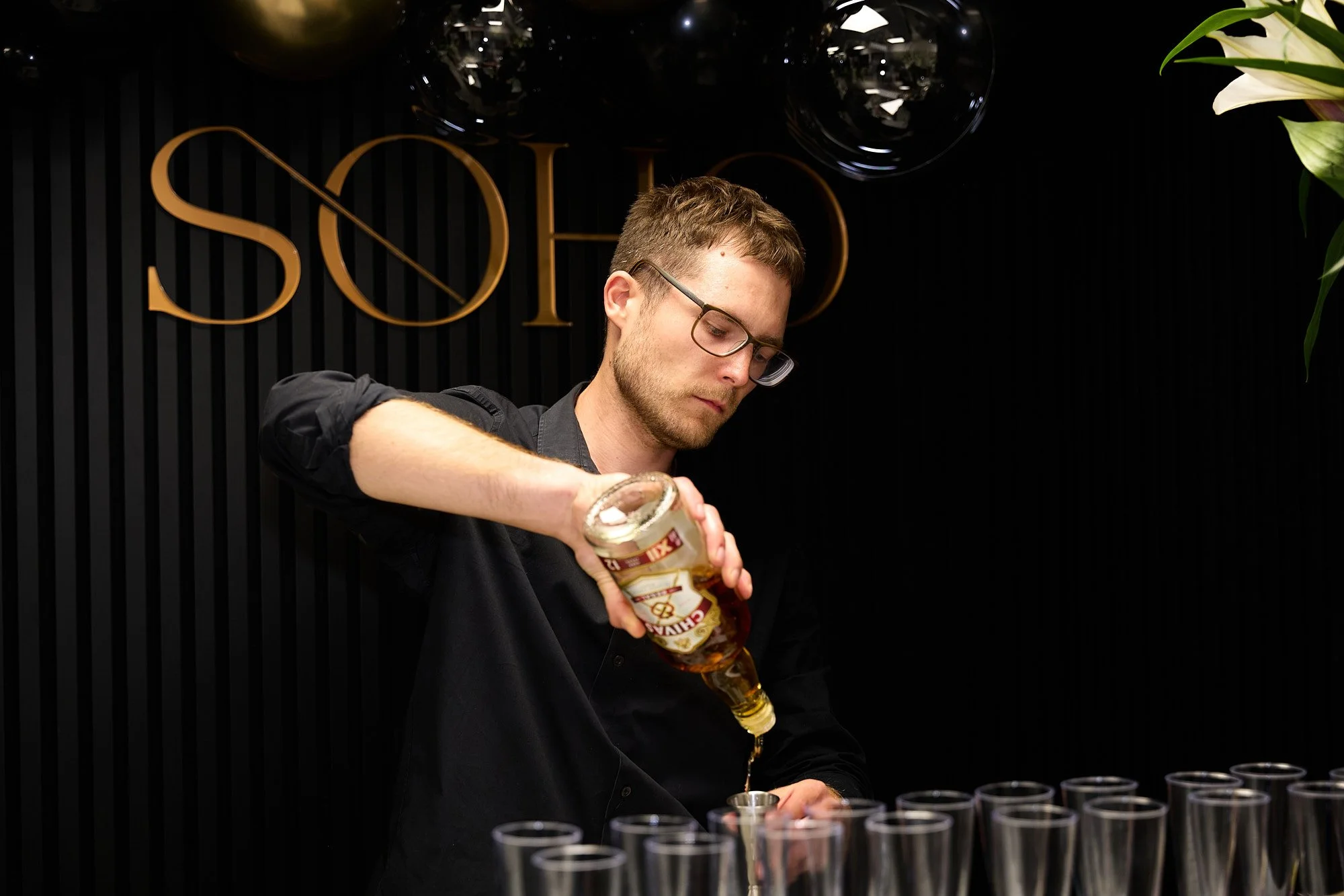 A bartender pouring beer from a can into a glass at a bar with black striped wall and a sign that says 'SOLD' in the background.