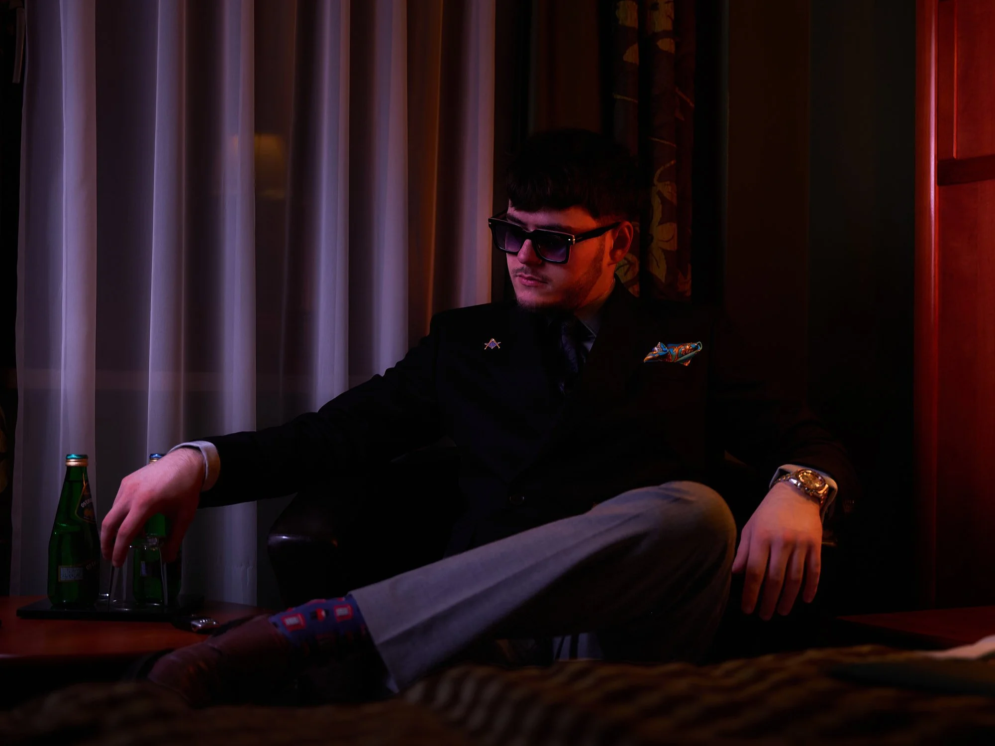 Young man dressed in black suit with colorful pocket square, wearing sunglasses, sitting in a dimly lit room, holding a bottle of sparkling water and a glass.