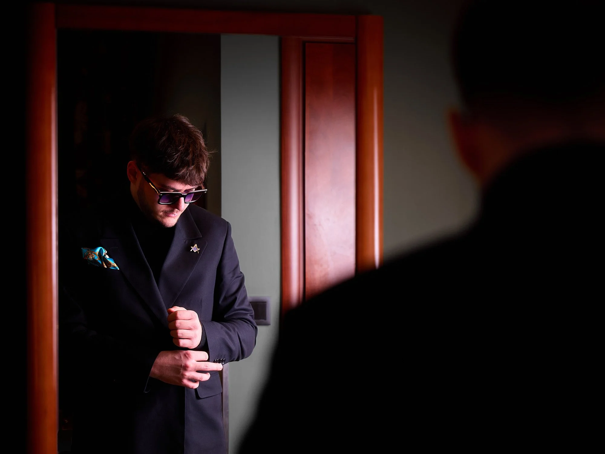 A man in a black suit and purple sunglasses adjusting his sleeve in front of a mirror, with a partial silhouette of another person in the foreground.