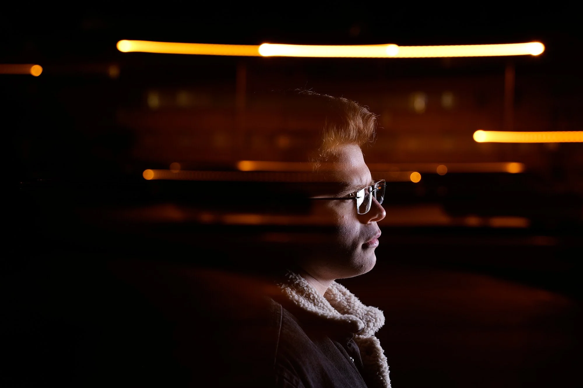 Side profile of a young man wearing glasses, looking ahead in a dark environment with blurred orange lights in the background.