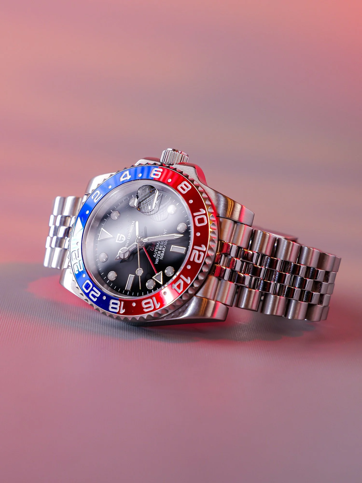 A wristwatch with a black dial, silver markers, and a red and blue bezel, lying on a pink gradient surface.