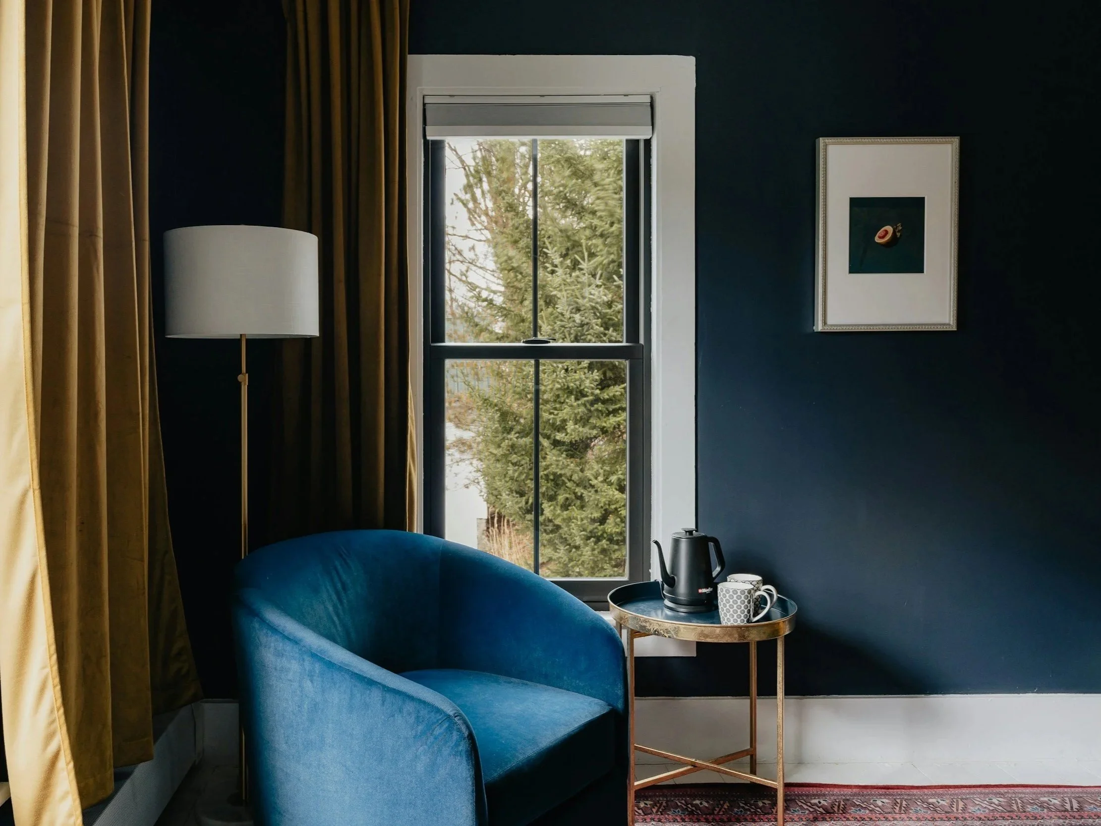 A cozy room corner with a dark blue wall, a blue velvet armchair, a tall white floor lamp, a window showing green trees outside, a small round side table with a black electric kettle and two cups, and a framed piece of art on the wall.