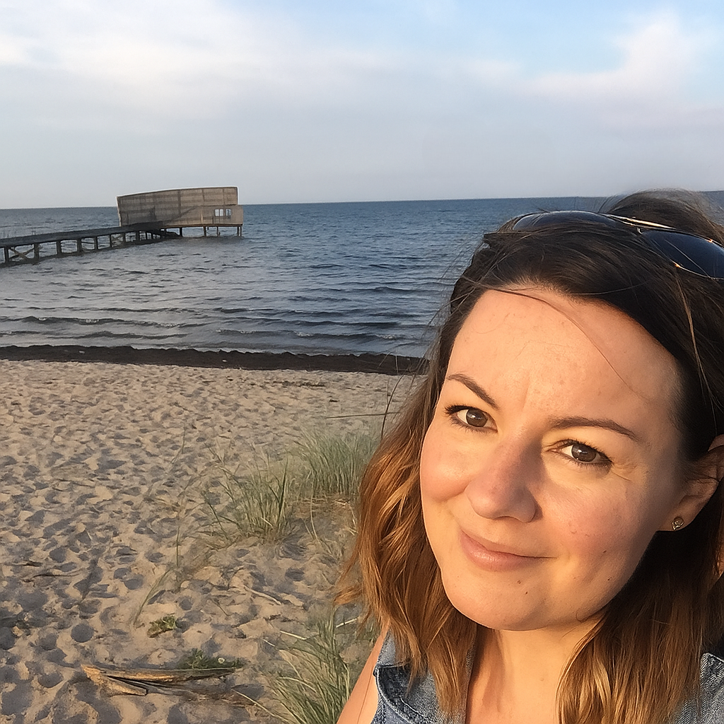 A young woman with brown hair and sunglasses on her head taking a selfie on a beach with sand, grasses, a pier, and the ocean in the background during sunset.
